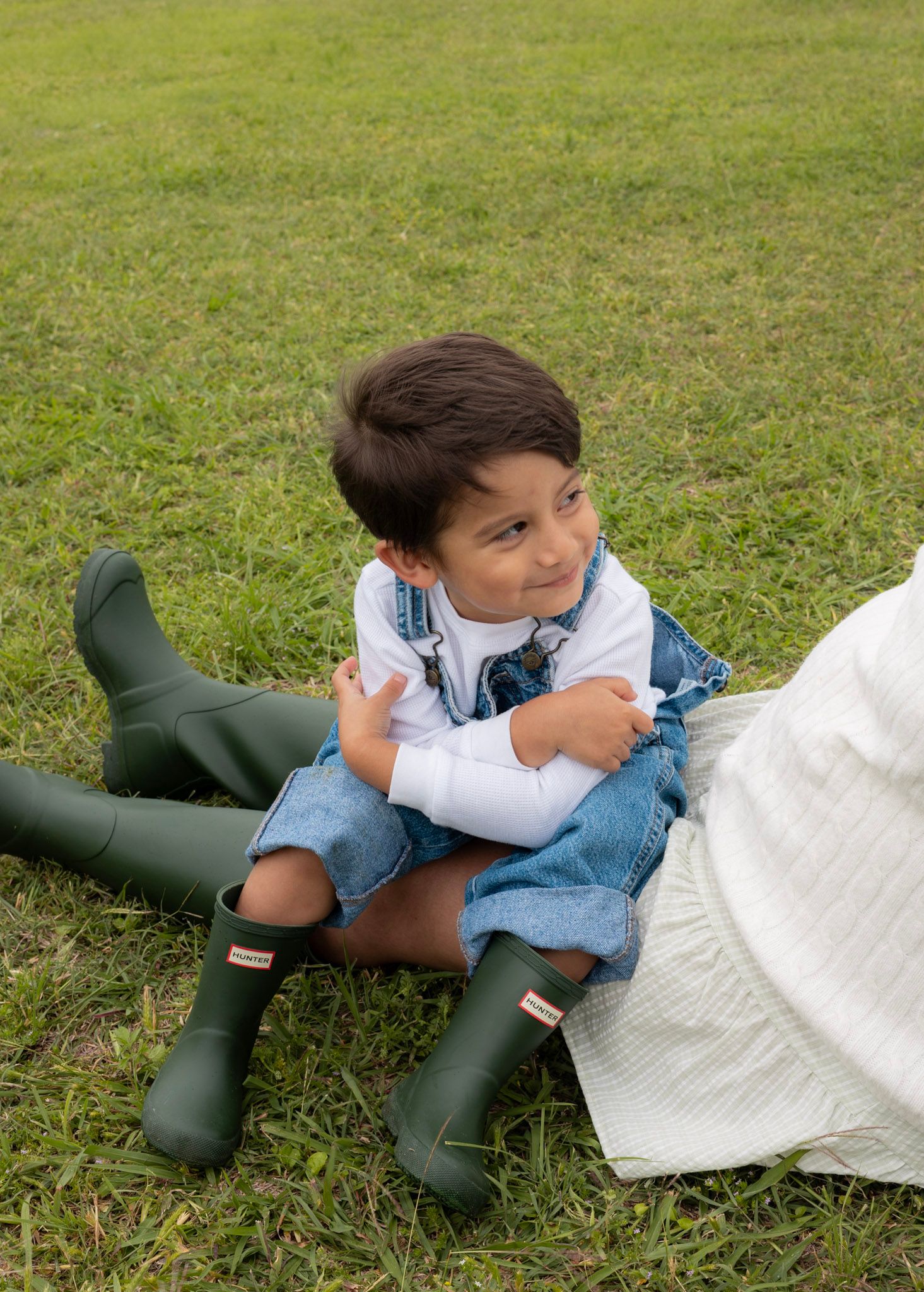 Young boy in denim overalls and green Hunter boots sitting on grass smiling.