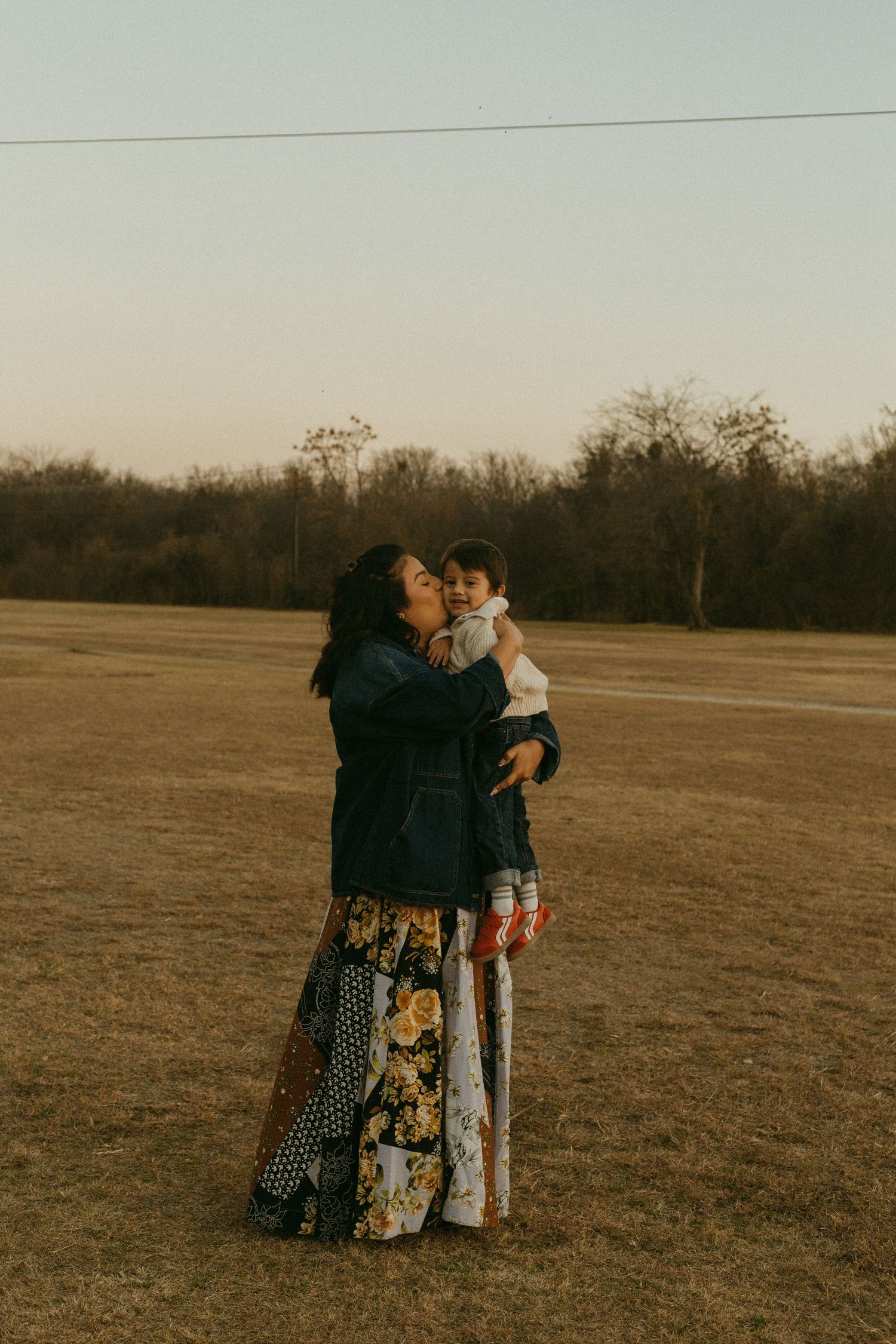 A mother kisses her baby while standing in an open field at golden hour, wearing a floral maxi skirt.