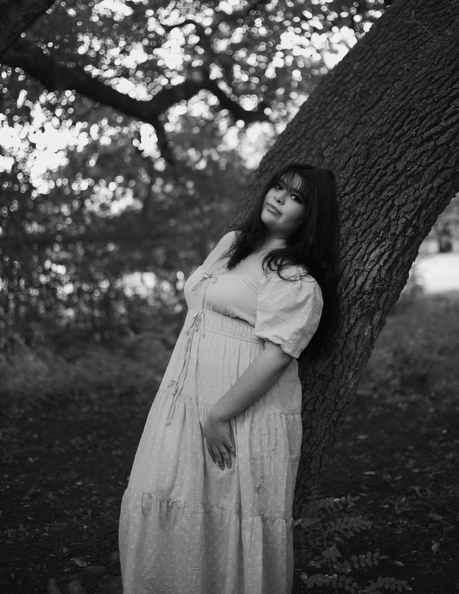 Black and white portrait of young woman in flowy dress leaning against a tree outdoors.