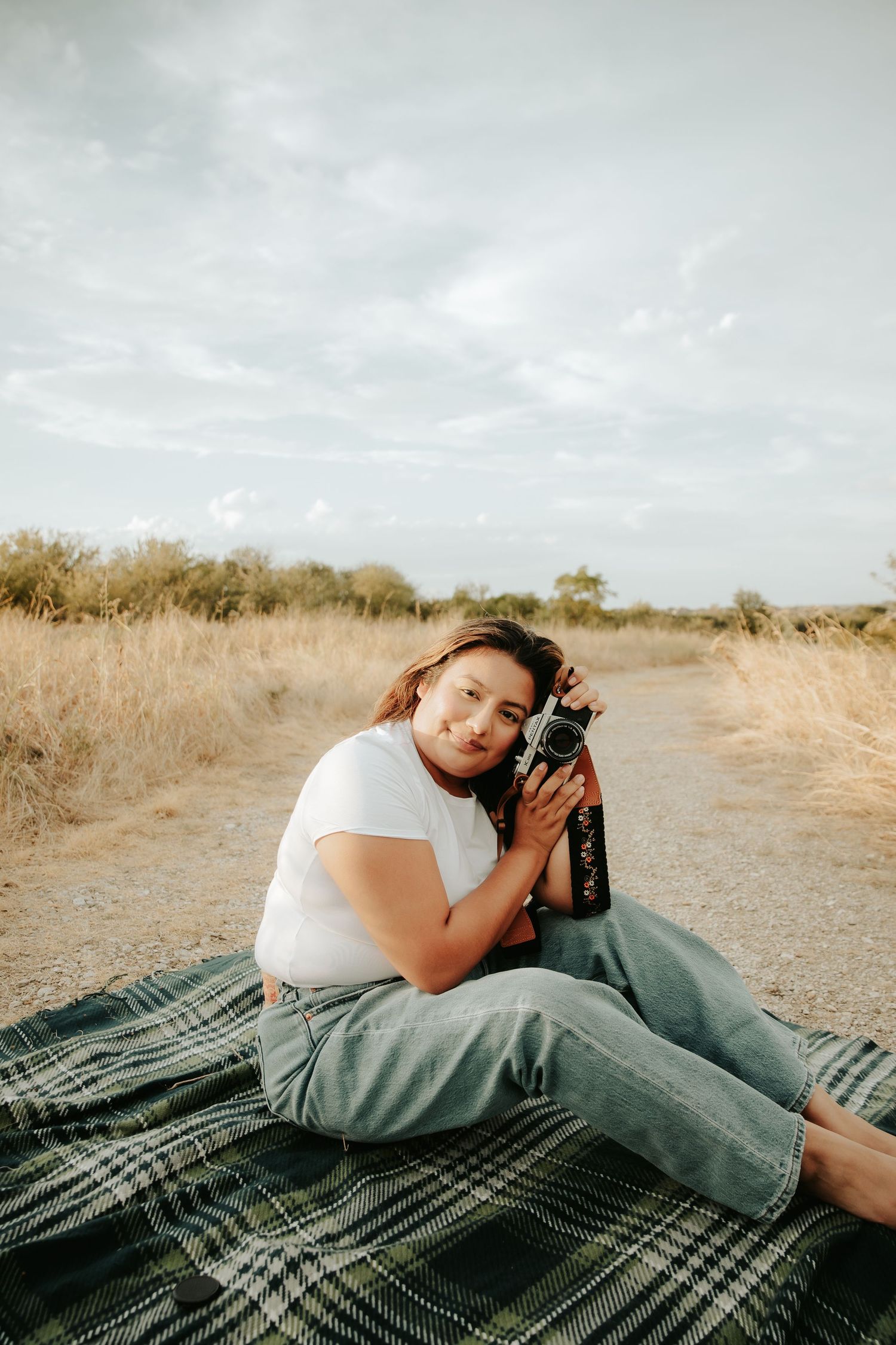 Woman in white tee and jeans sits on plaid blanket outdoors, holding vintage camera against her cheek in golden field.