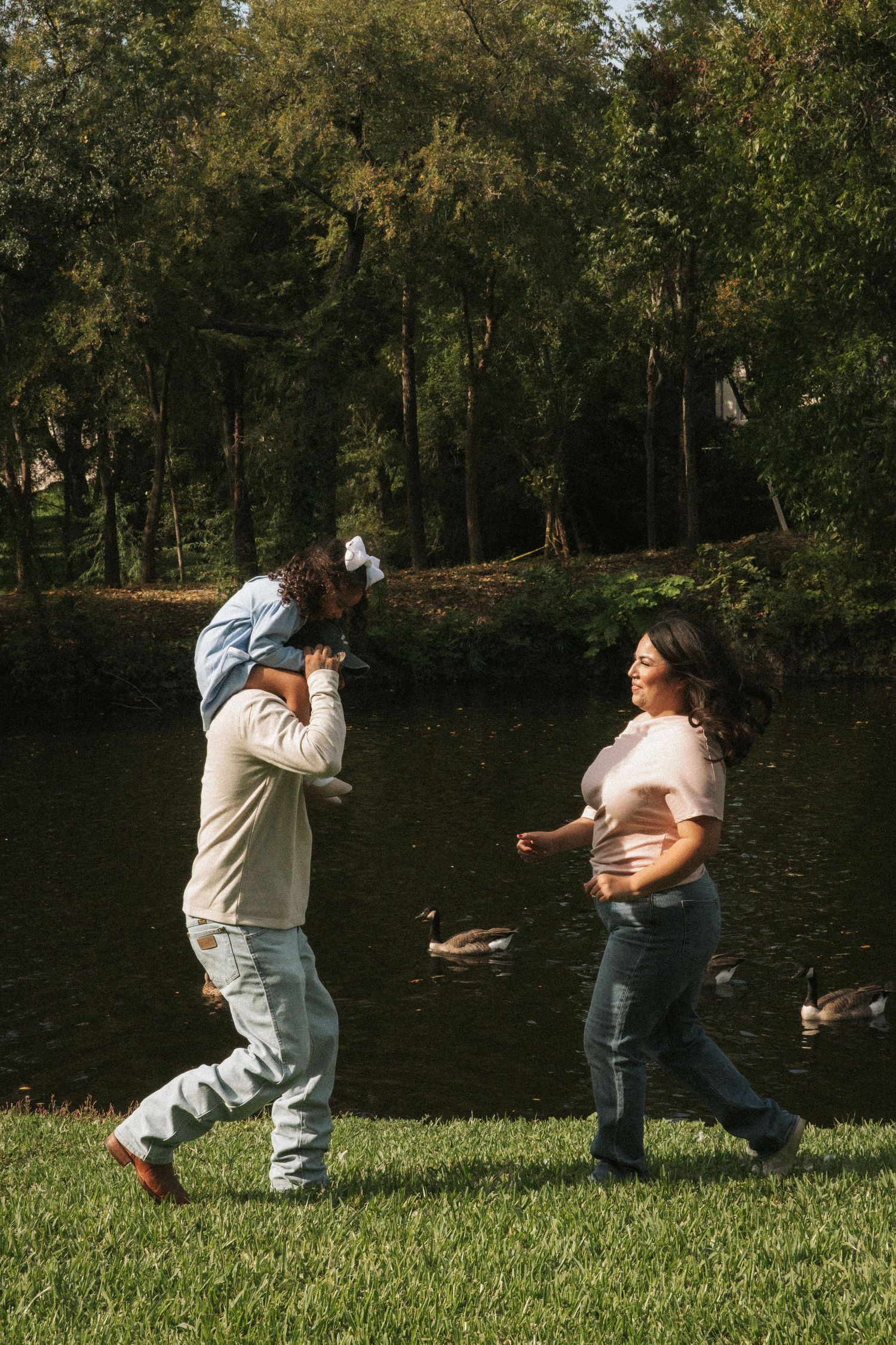 Family walking by a pond in a park, father carrying daughter on shoulders while mother walks alongside them.