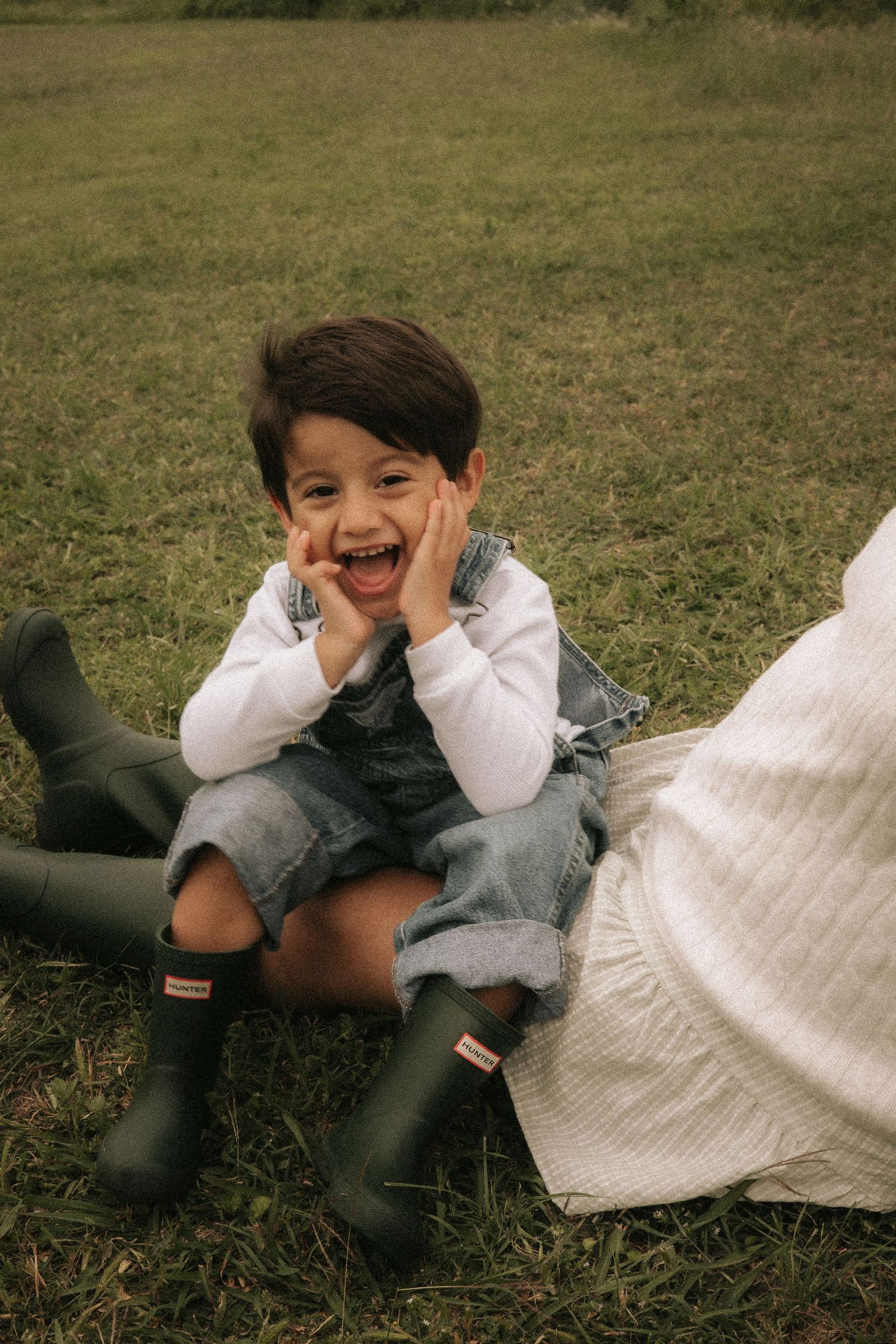 A joyful young child sitting on grass wearing denim overalls, white shirt, and black Hunter rain boots, laughing with hands on cheeks.
