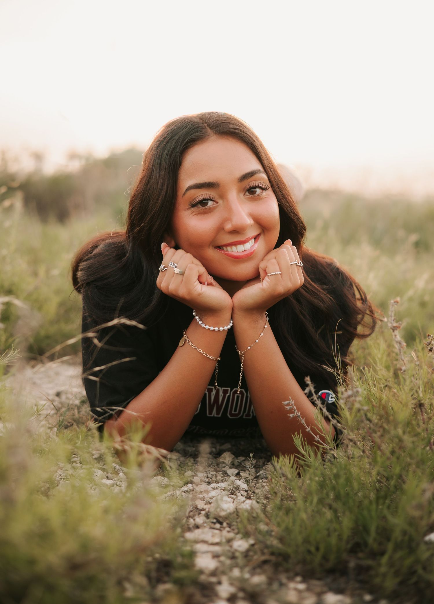 Young woman lying in a grassy field, smiling with hands under chin, wearing a black sweatshirt and bracelets.
