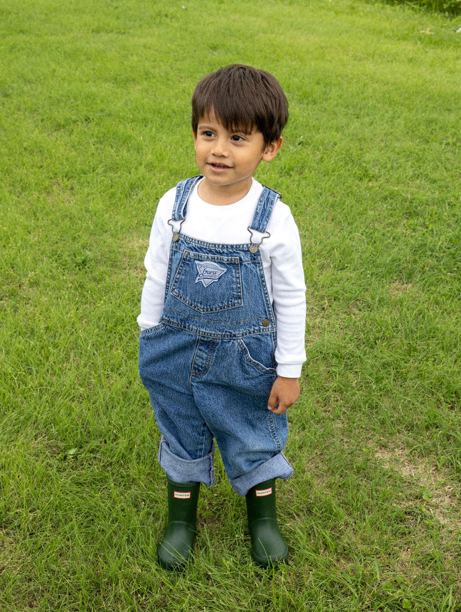 Adorable toddler boy in denim overalls and green Hunter boots standing on green lawn.