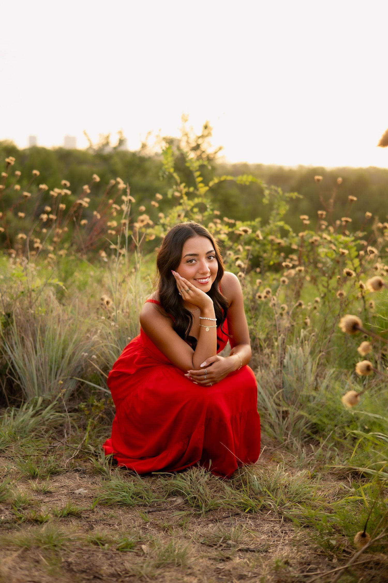 Smiling young woman in red dress seated in a sunlit wildflower meadow at golden hour.