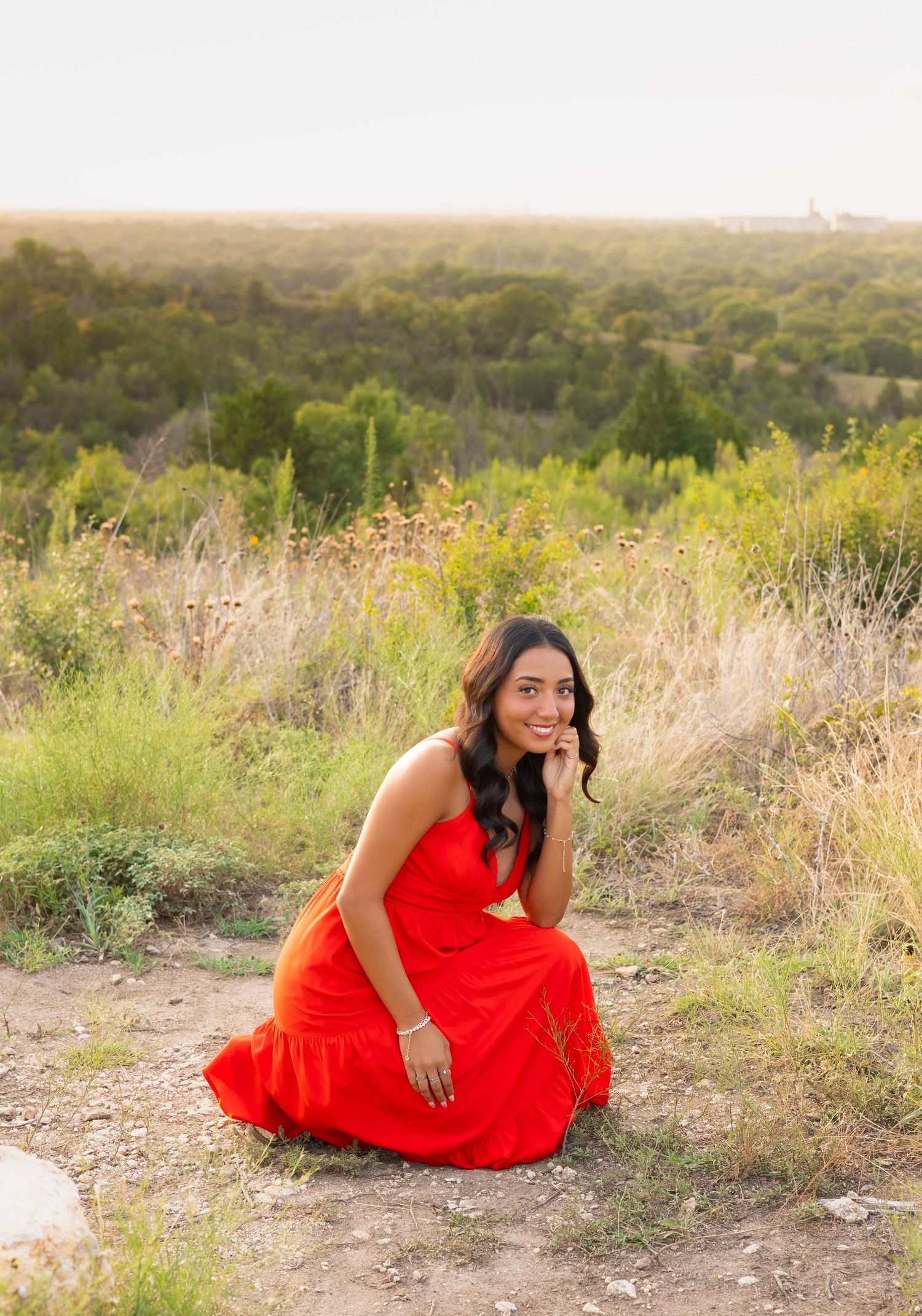 Smiling woman in red dress crouching on a dirt path with scenic green landscape behind her.