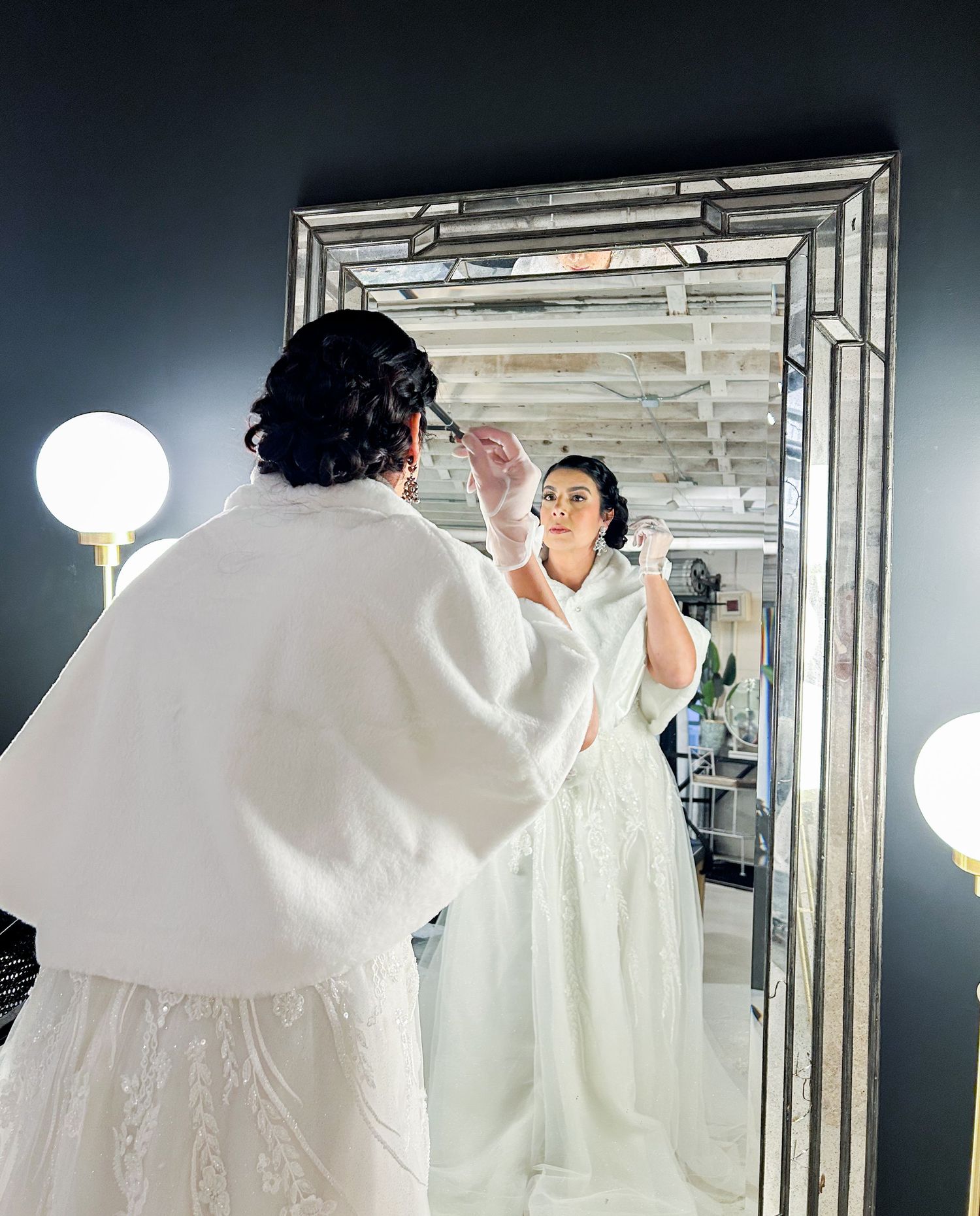 Bride in white wedding dress and bolero jacket admiring her reflection in a large ornate mirror while getting ready.