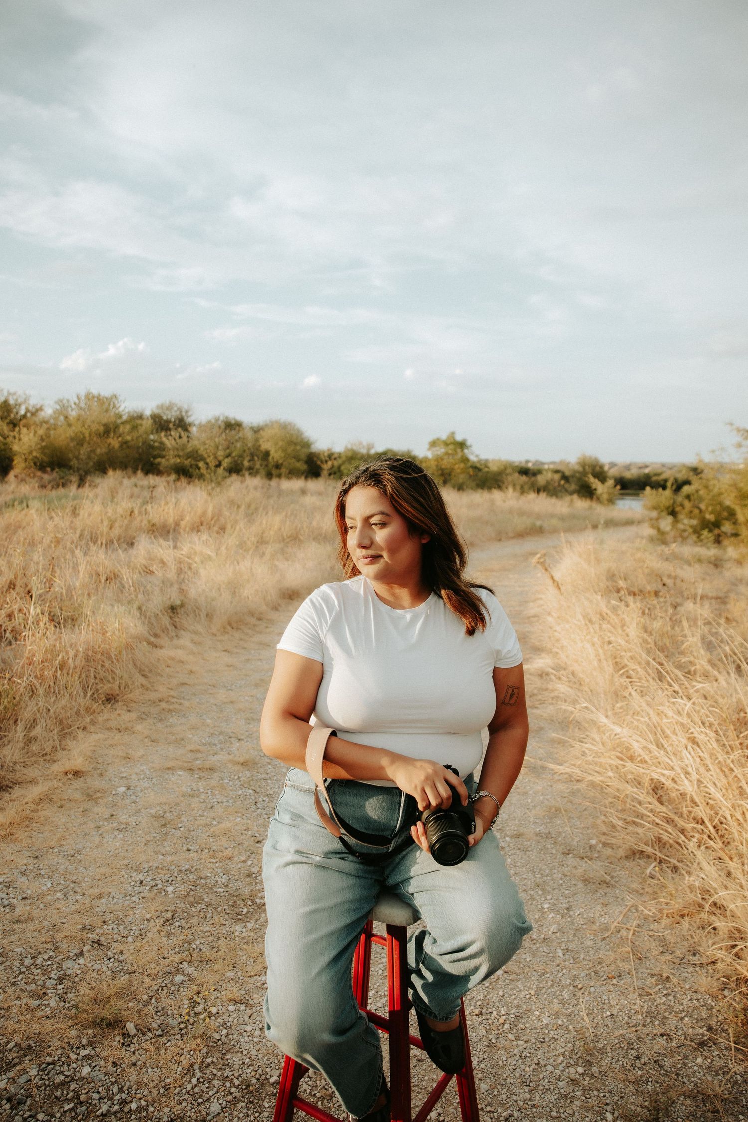 Woman in white t-shirt and jeans sitting outdoors on red stool holding camera in golden field.