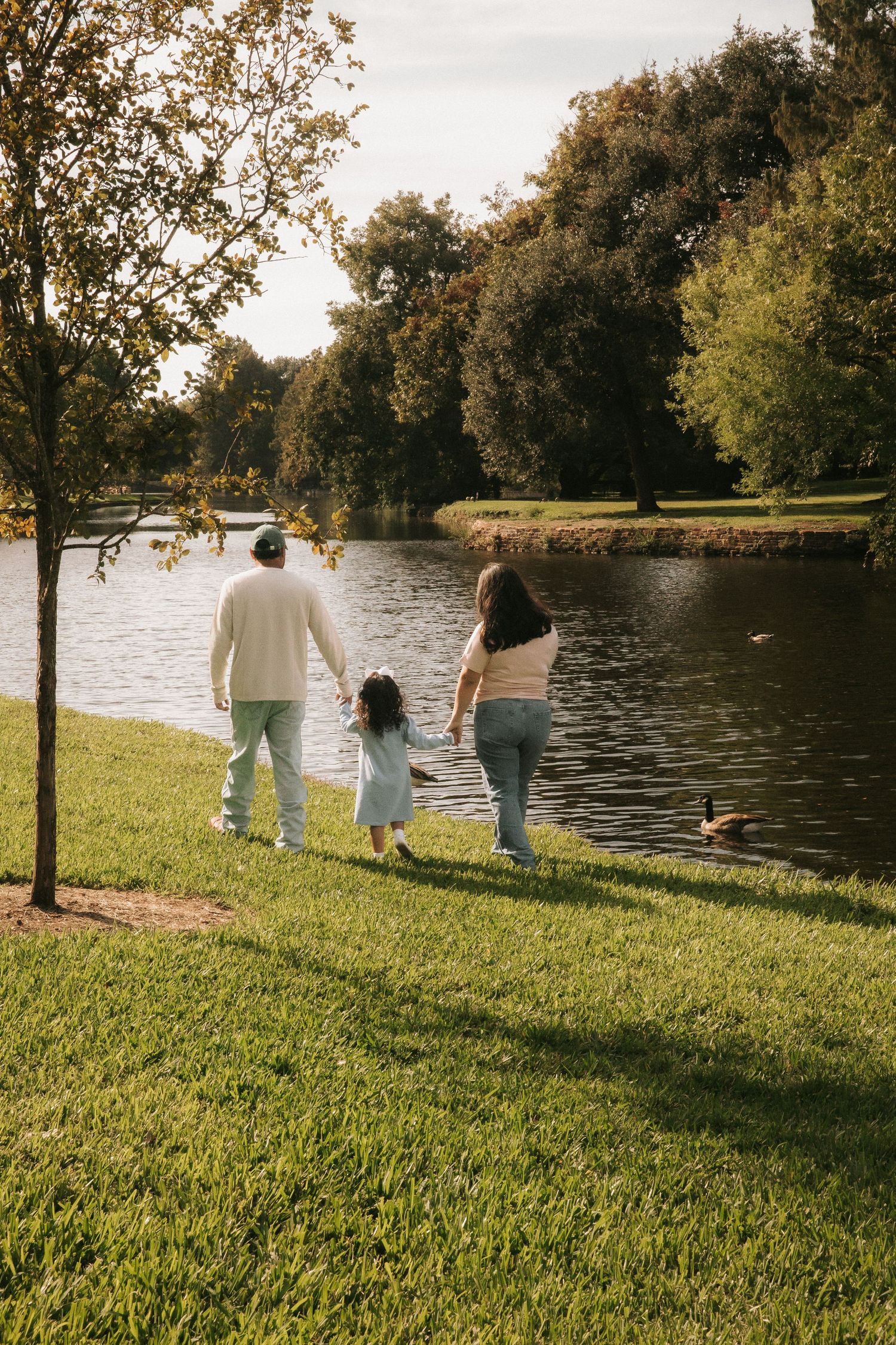 Family of three holding hands walking toward a serene park lake surrounded by lush green trees on a sunny day.