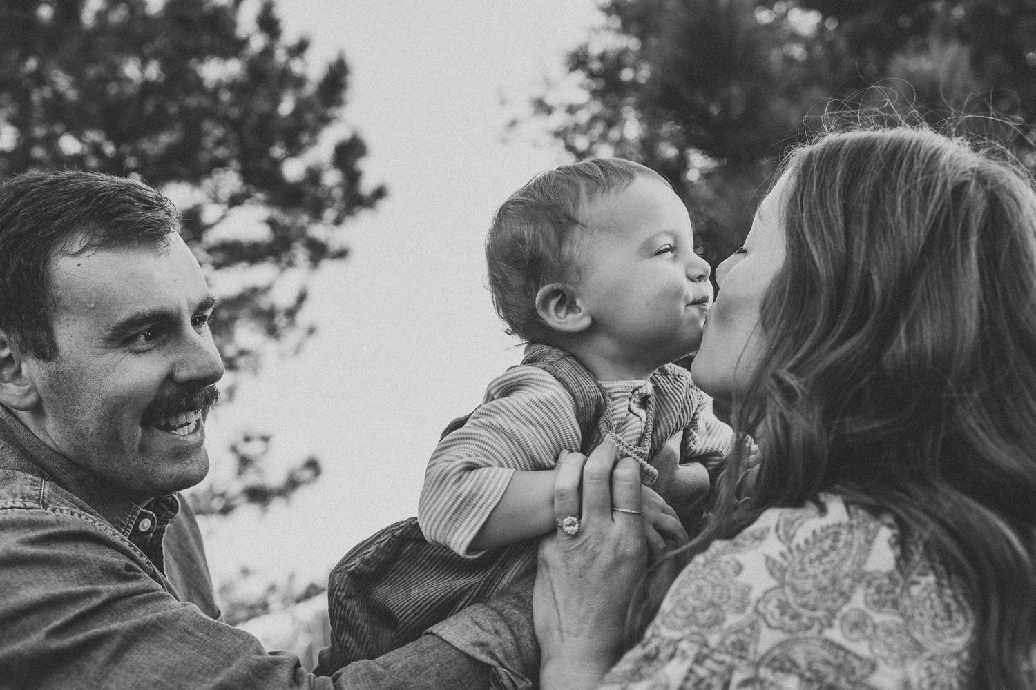 Black and white photo of smiling parents holding their baby outdoors, mother kissing infant cheek near trees.