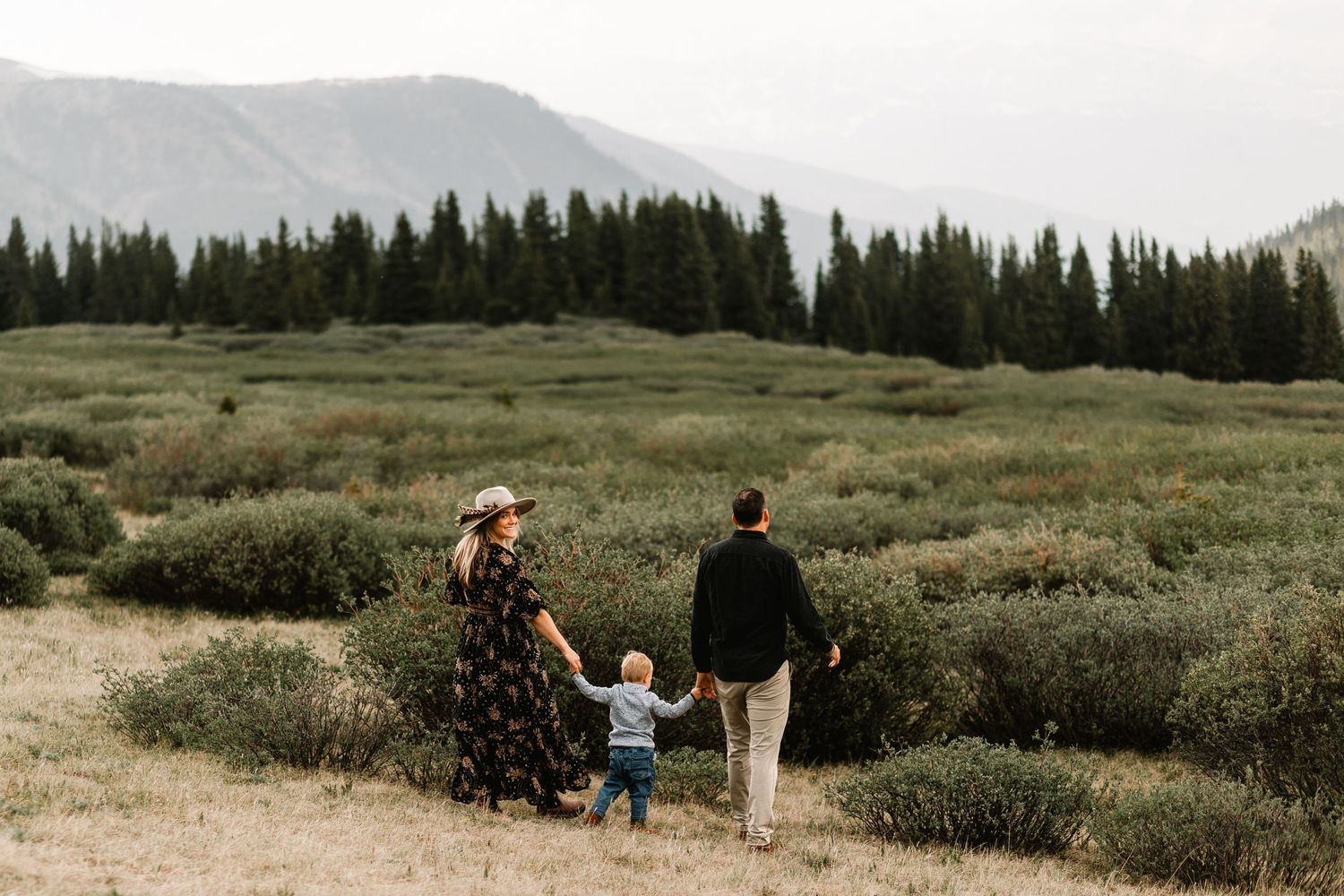 Family of three walking through a lush mountain meadow with evergreen forest backdrop.