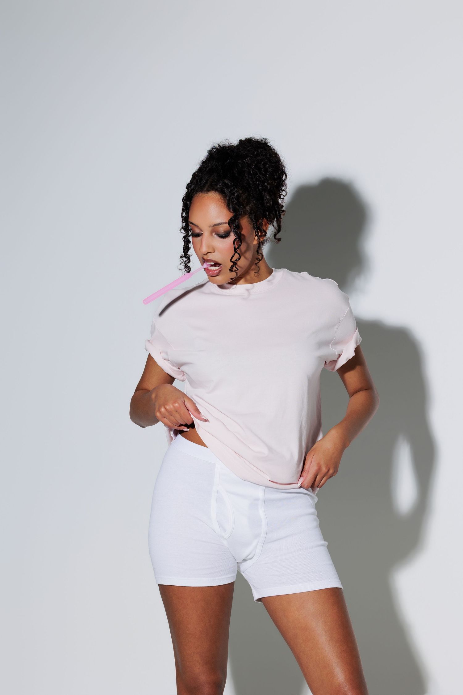 Woman in pink t-shirt and white shorts posing with hands on hips, holding a pink straw against a grey background.