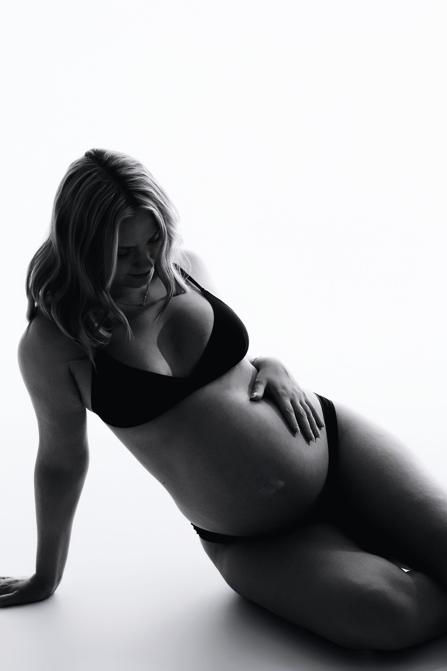 Black and white photo of a pregnant woman in lingerie sitting on the floor touching her belly.
