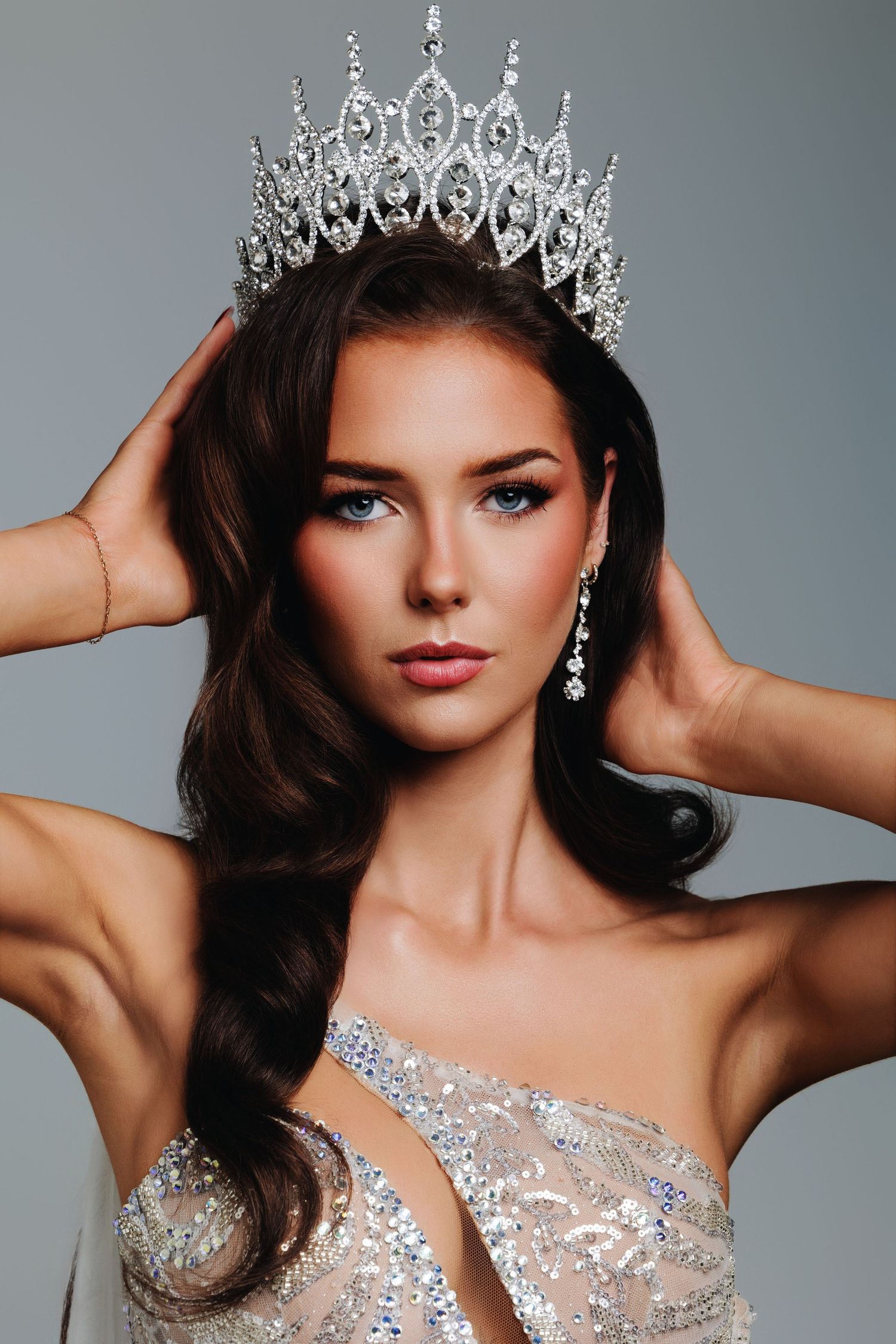 Young woman wearing a sparkling crystal crown and sequined gown poses elegantly with hands raised to her tiara.