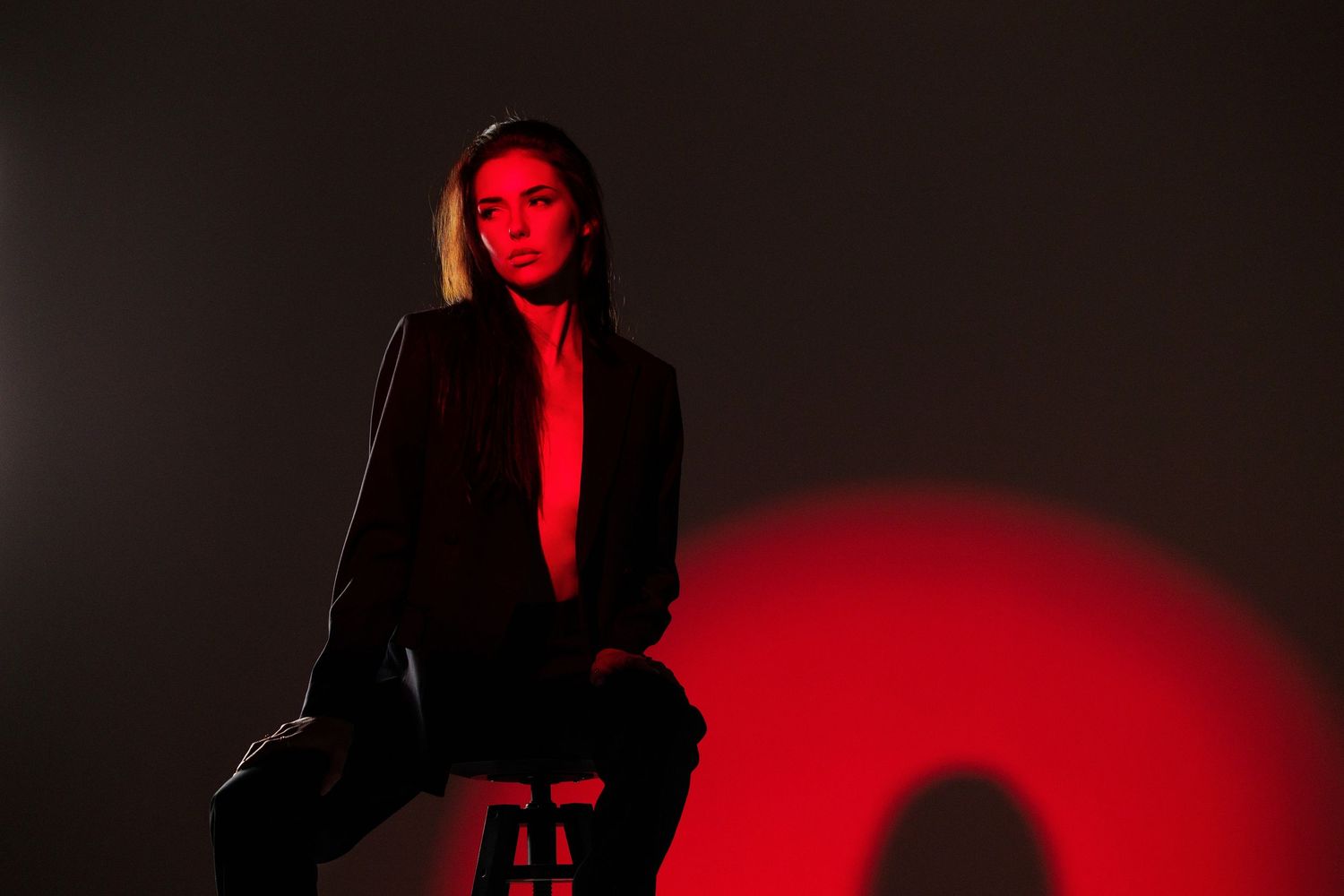 Woman in black blazer poses on stool under dramatic red spotlight against dark studio background.