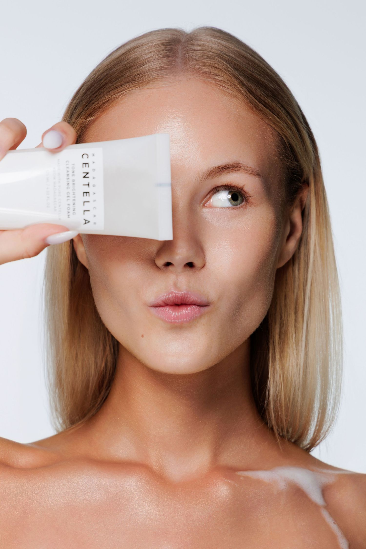 Blonde woman covering one eye with Centella facial cleanser tube against white background.