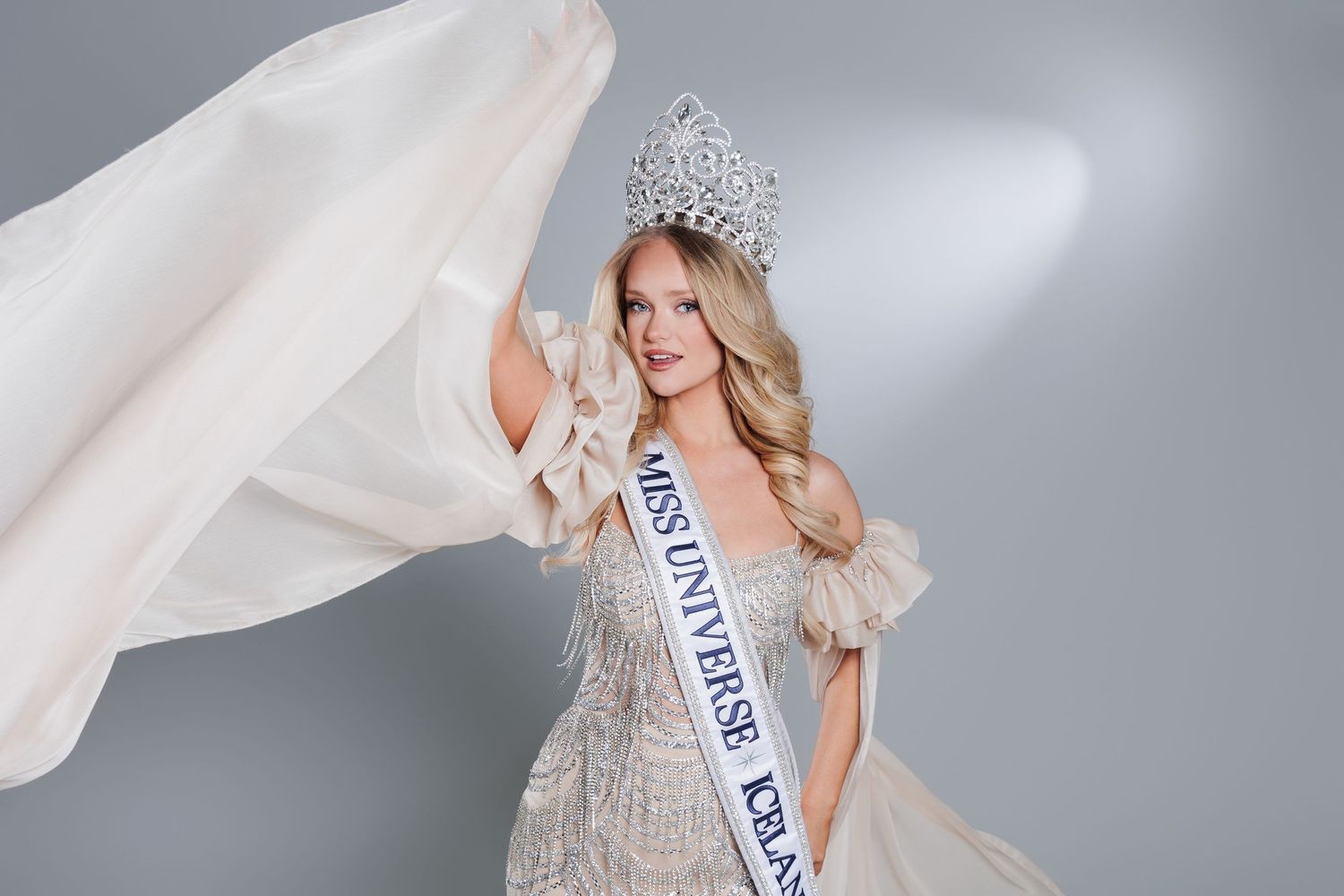 Miss Universe contestant in a sparkling gown and crown poses elegantly with flowing white fabric.