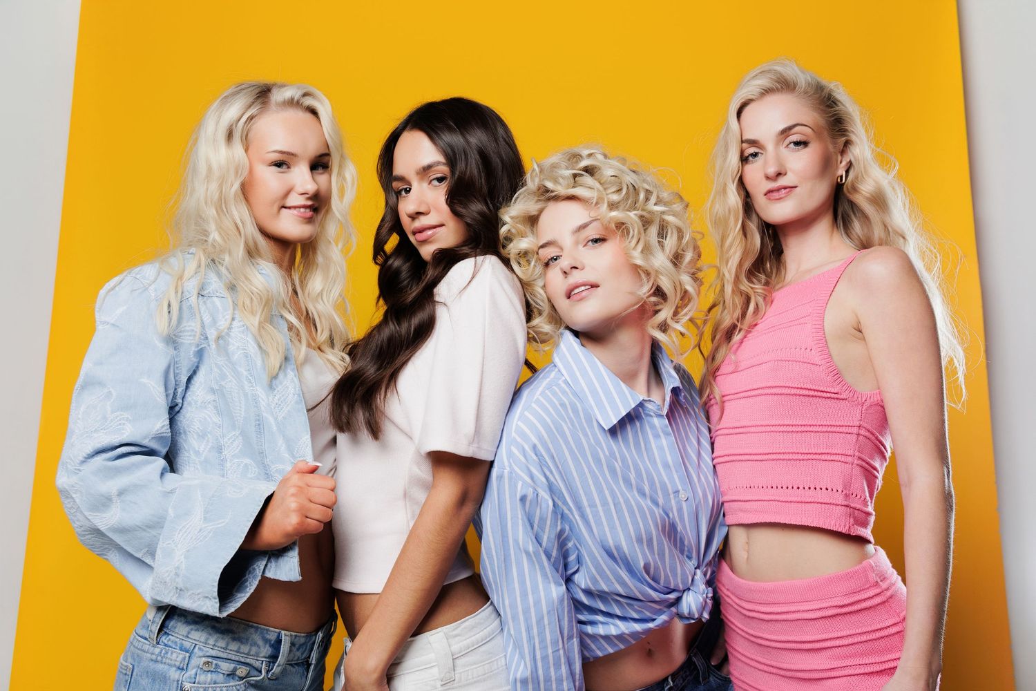 Four young women posing together in casual outfits against a bright yellow background.