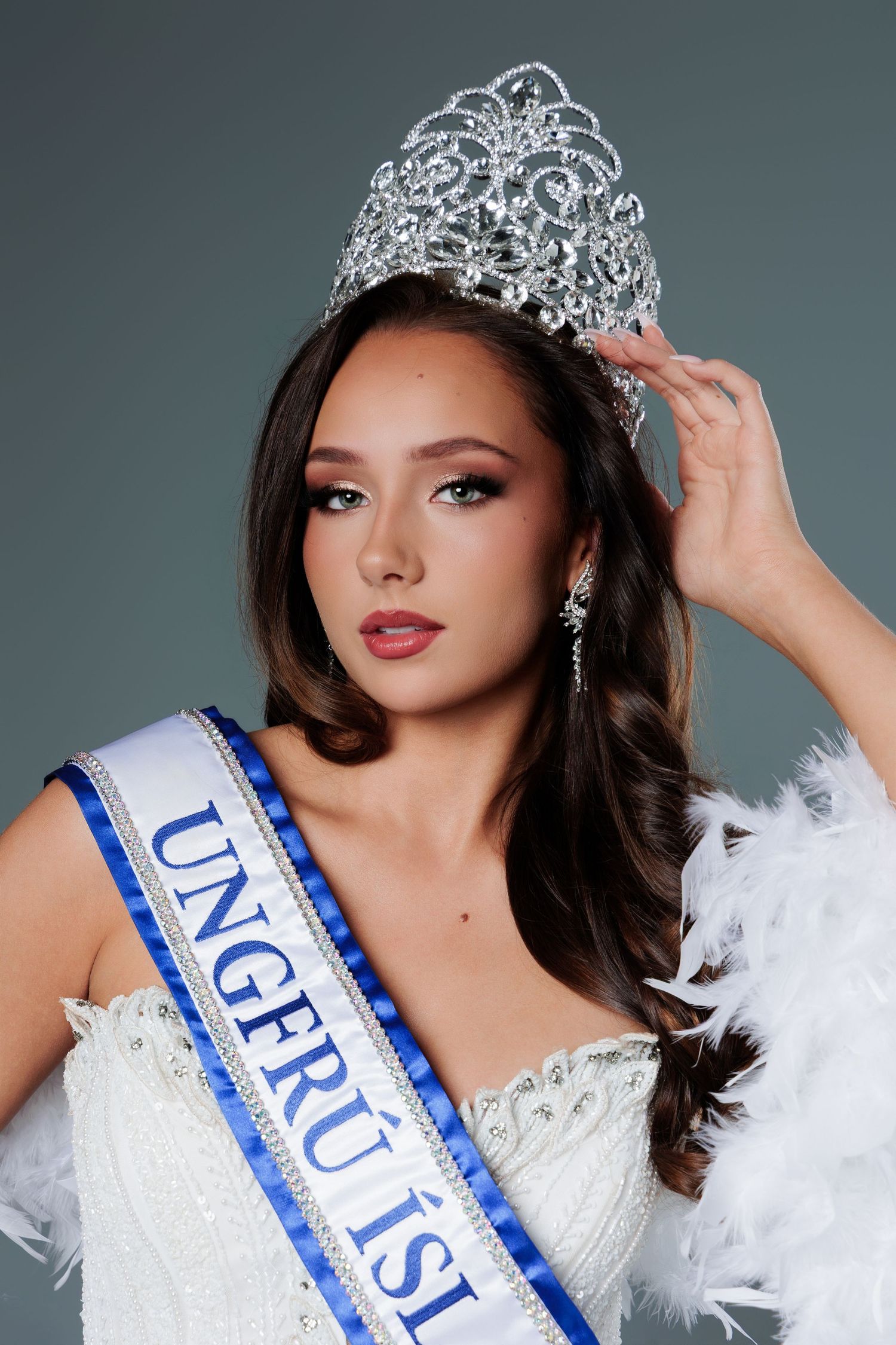 Young woman wearing a pageant crown and blue sash reading Ungfrú Ísland in a white gown with feather trim.