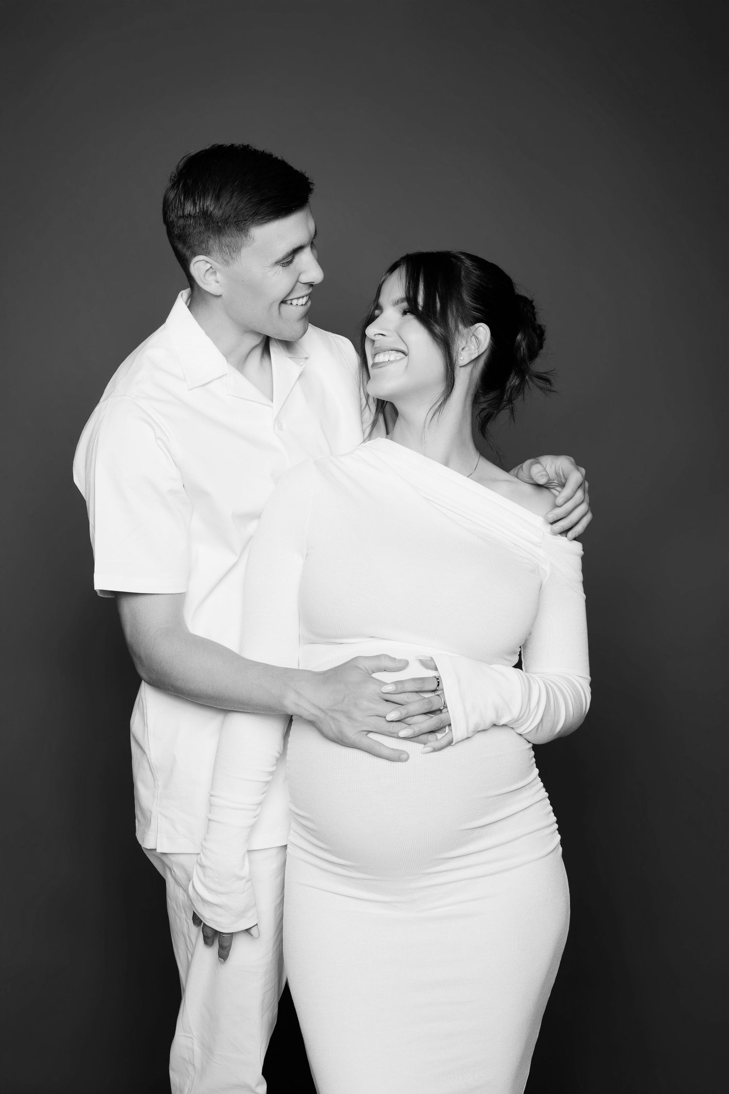 Black and white photo of smiling couple, man embracing pregnant woman in white dress in studio.