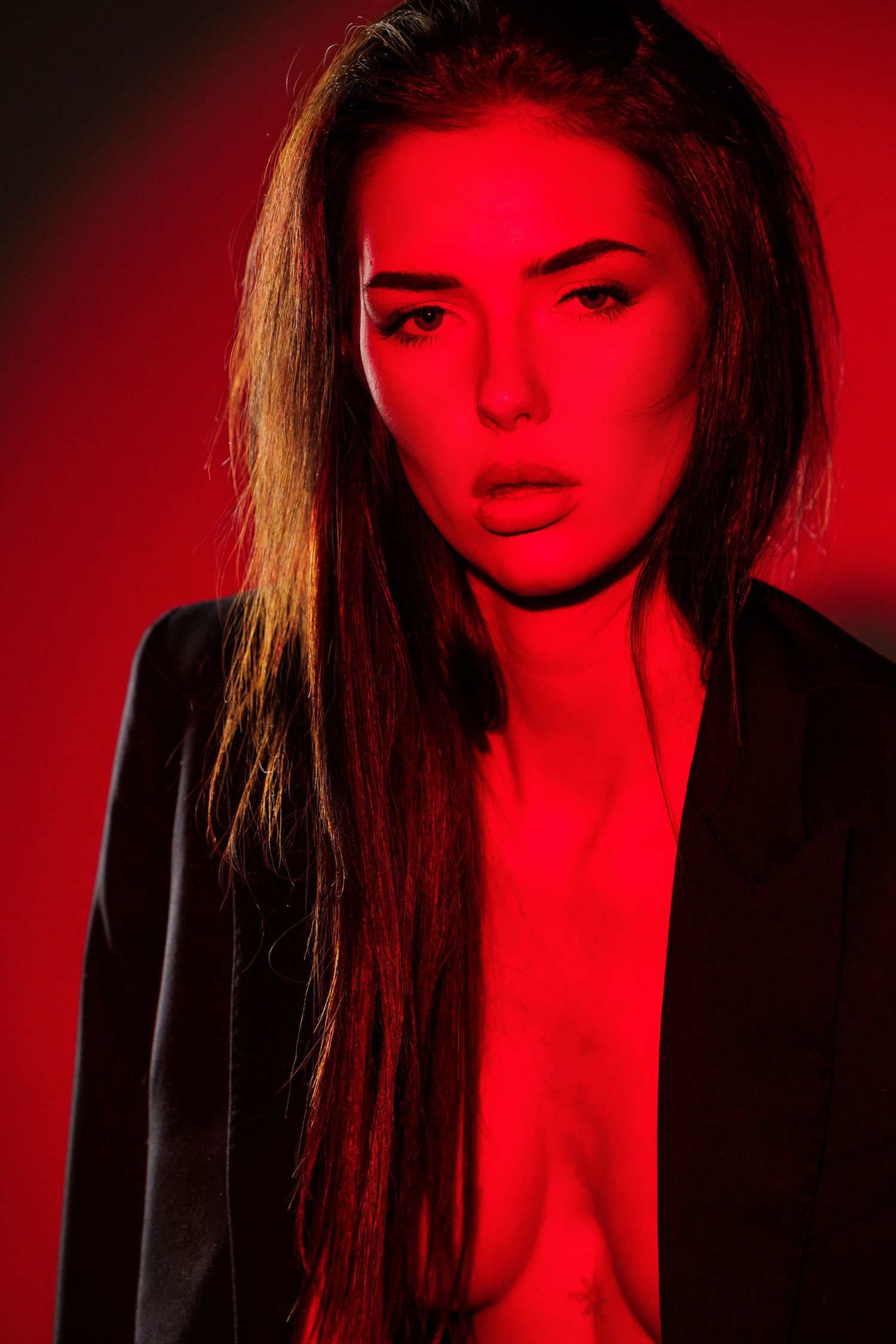 Woman with long hair in black blazer posed against dramatic red-lit background in moody portrait photography.