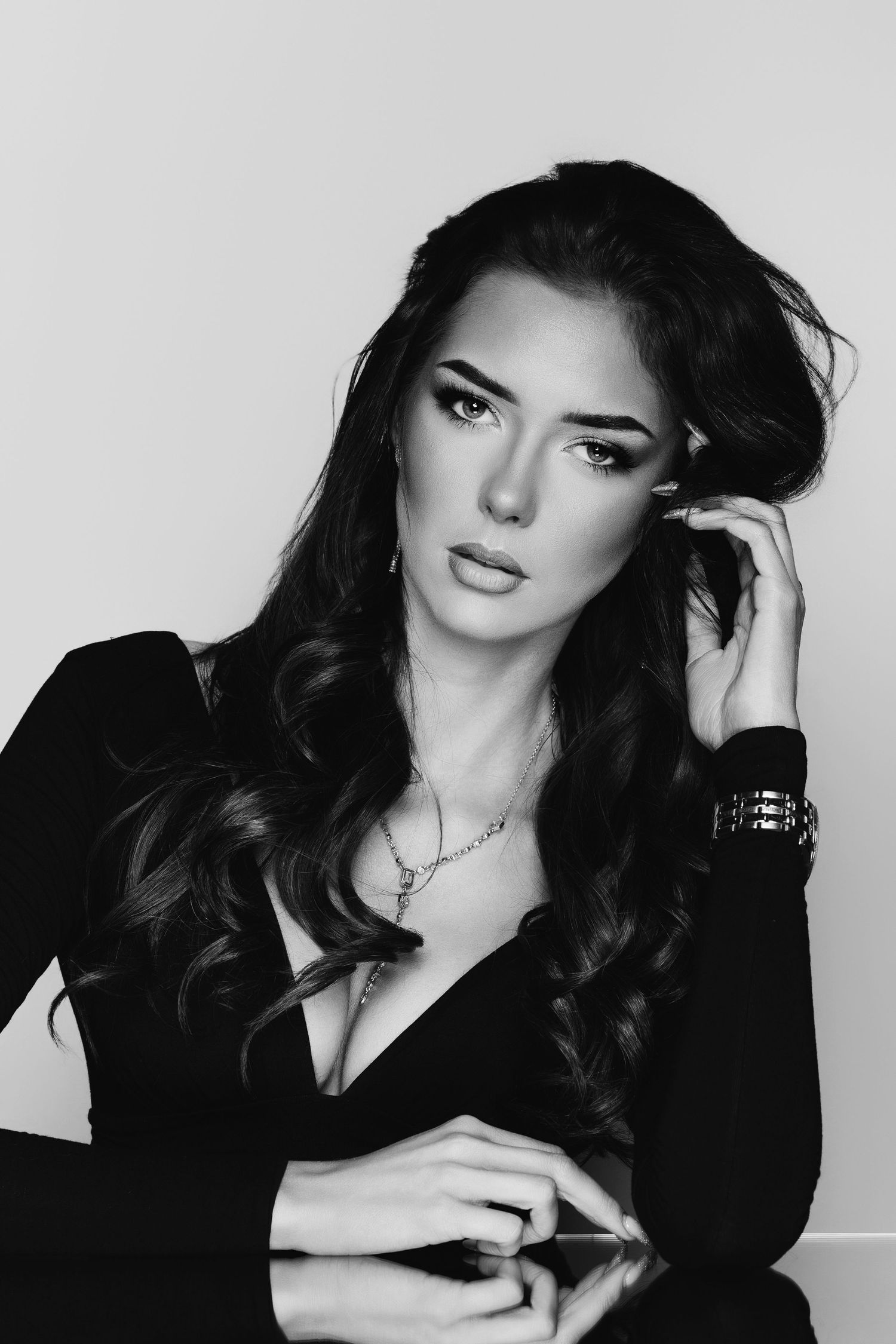 Black and white portrait of a brunette woman in a black dress with jewelry, posing elegantly at a reflective table.