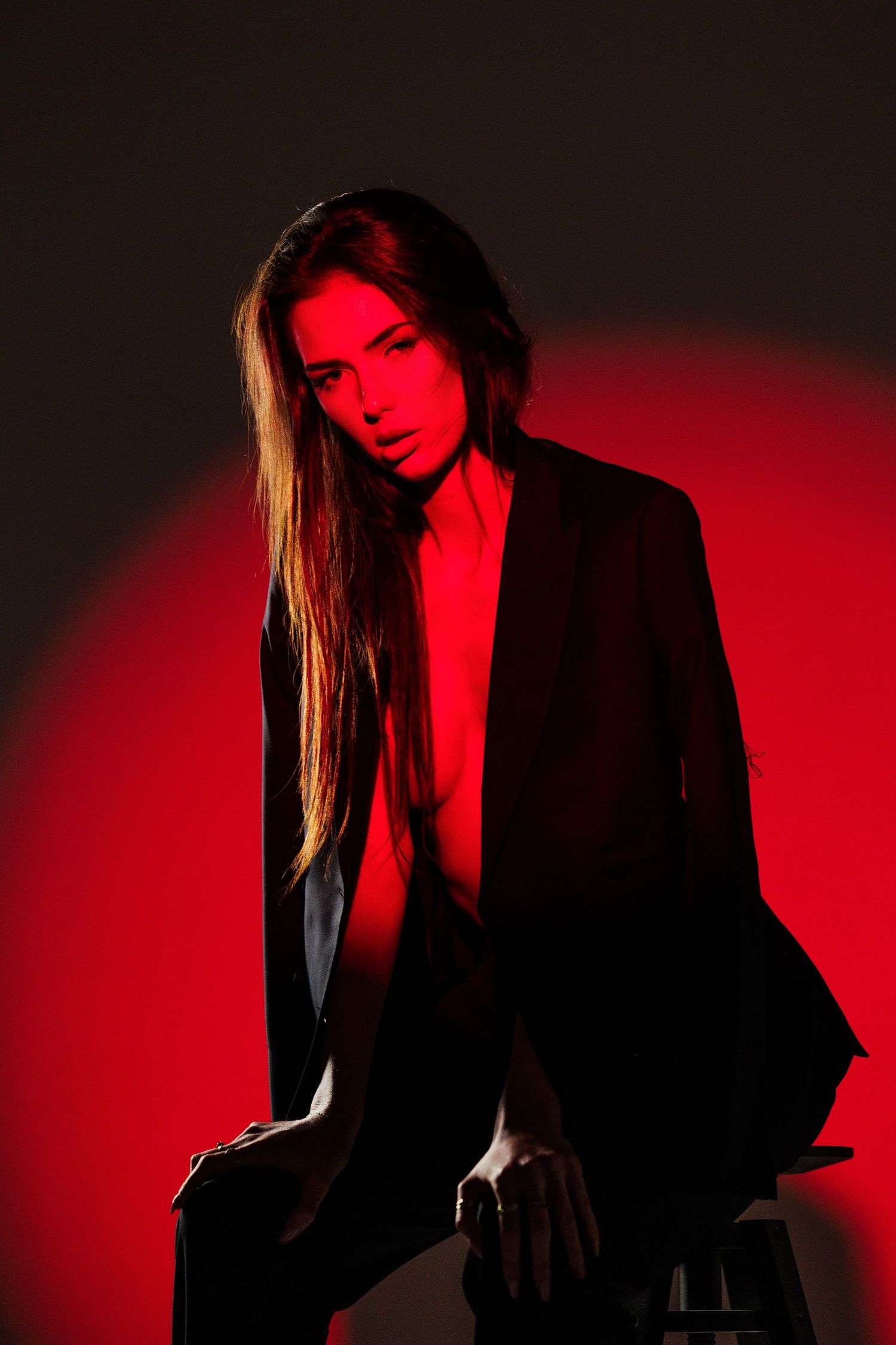 A woman in a black blazer poses dramatically under red studio lighting against a dark background.