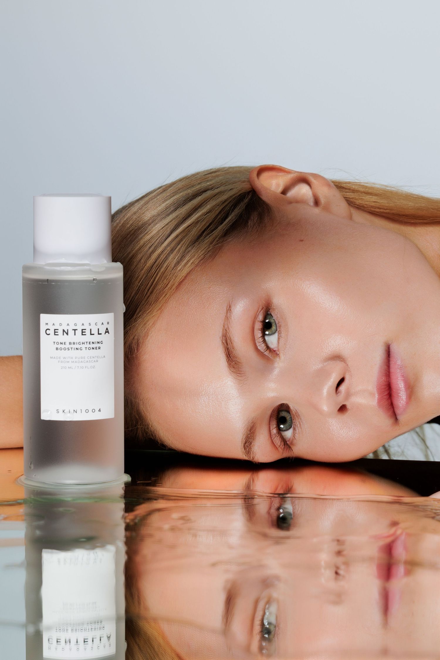 Young woman rests her face on a reflective surface beside Skintoca Centella toning exfoliating toner bottle.