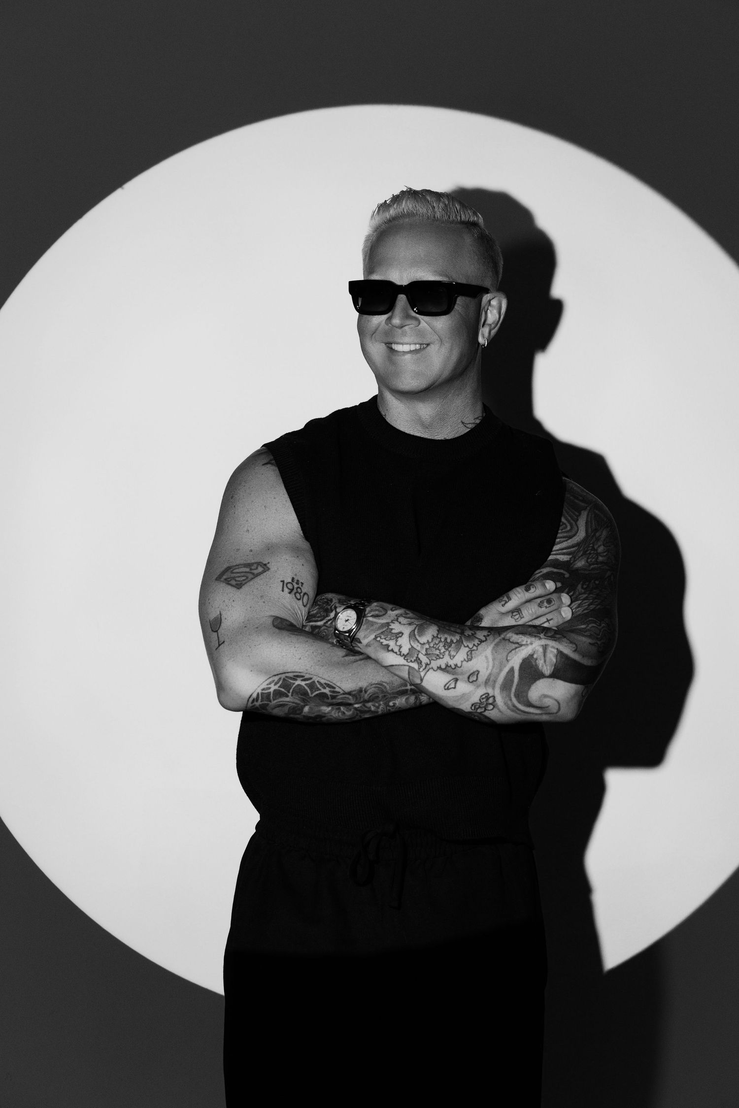 Black and white photo of tattooed man in sunglasses, arms crossed, smiling, wearing sleeveless black outfit.