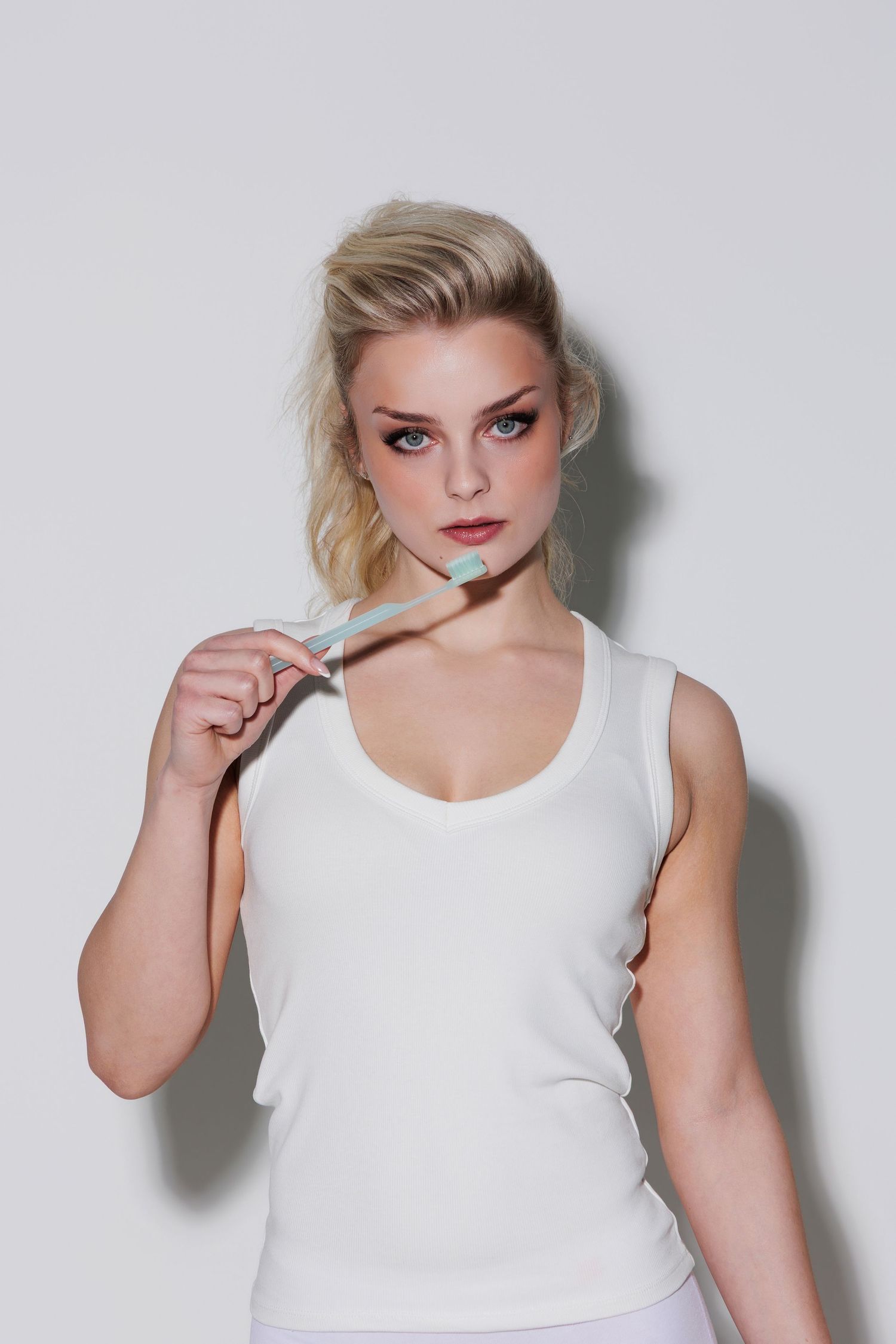 Blonde woman in white tank top holding a toothbrush, looking at camera against light background.