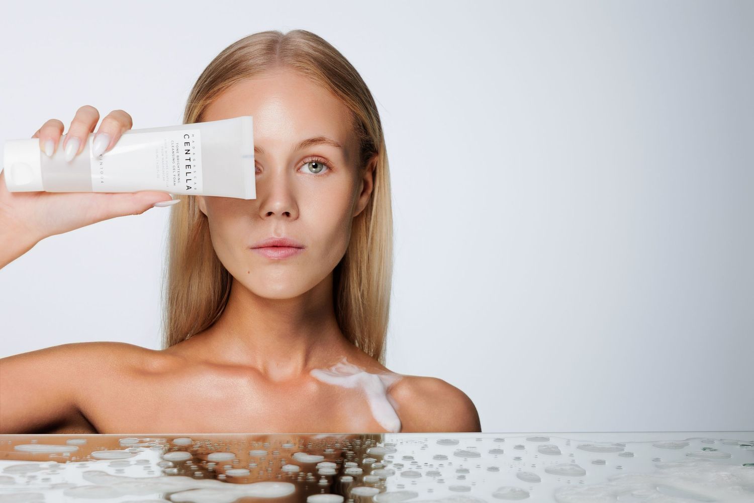 Woman holding white skincare bottle near face, emerging from water, showcasing hydrating beauty product on light background.