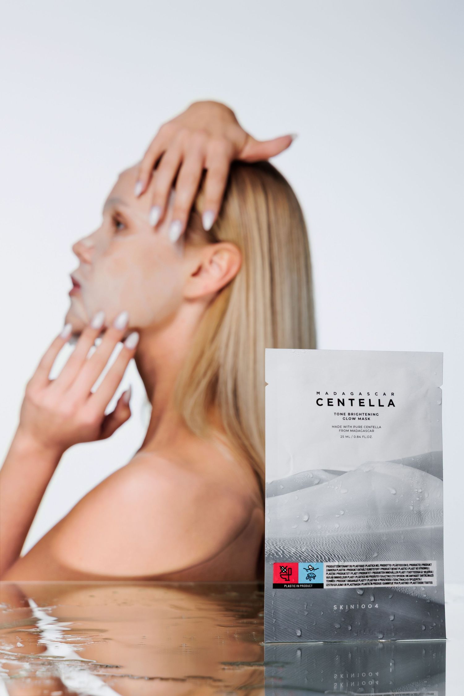 Blonde woman applying Madecassol Centella face mask sheet in water, with product packaging in foreground.