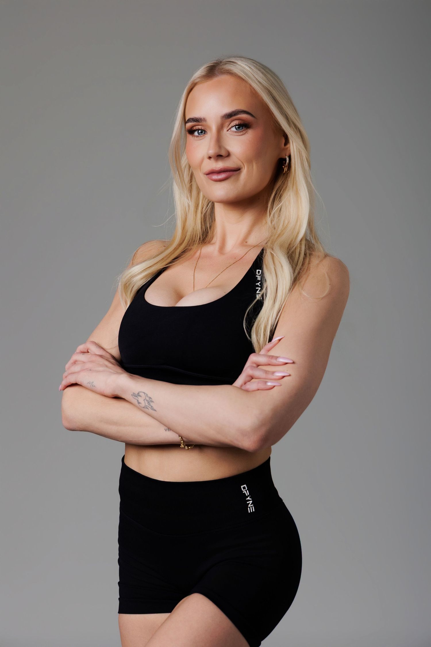 Blonde woman in black sports bra and shorts posing with arms crossed against gray background.
