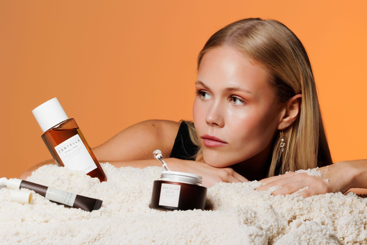 Woman posing with luxury skincare products including serum, jar, and bottle against an orange background.