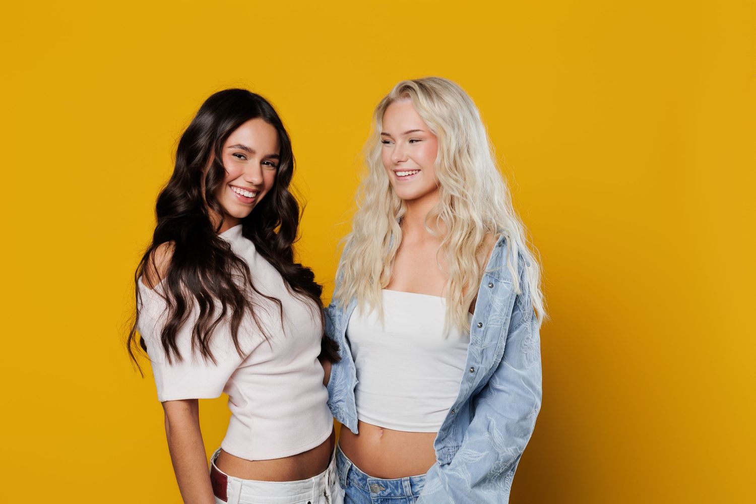 Two smiling women posing together against a bright yellow background, one brunette and one blonde, wearing casual crop tops.