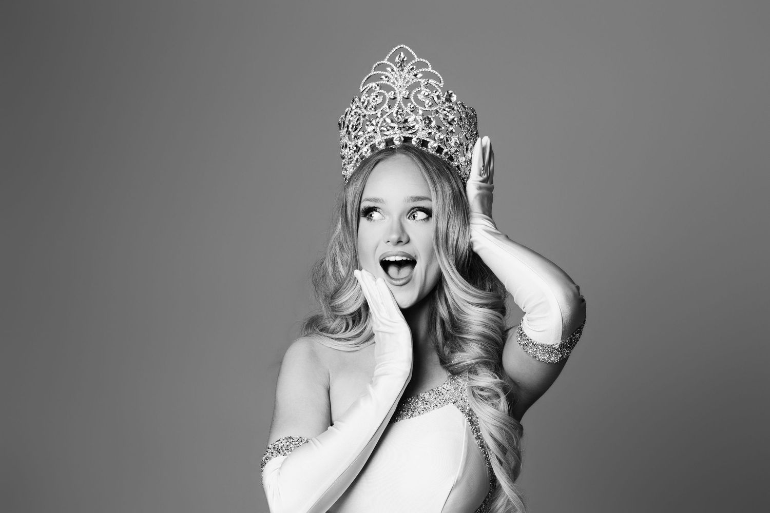 Excited young woman wearing a sparkling tiara crown, posing with hands on face in black and white photo.