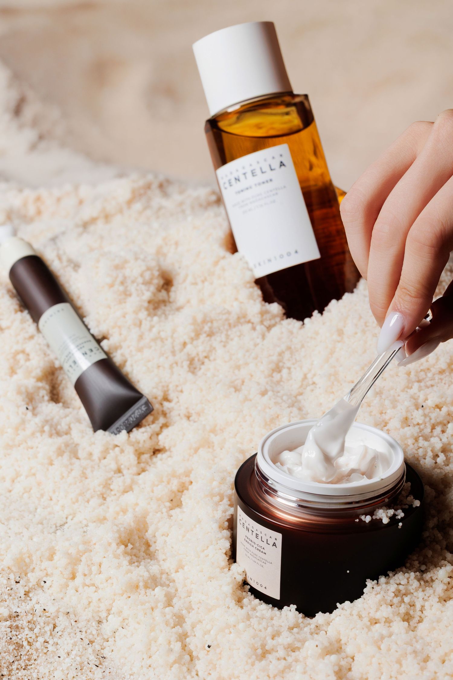 Centella skincare products arranged in white sand, featuring a cream jar, toner bottle, and serum with a dropper applicator.