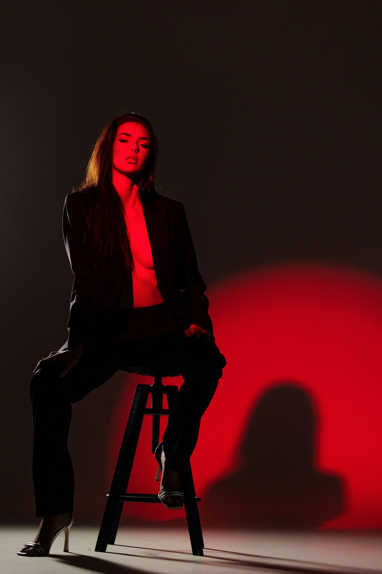 Woman in black suit sitting on stool under dramatic red studio lighting against dark background.