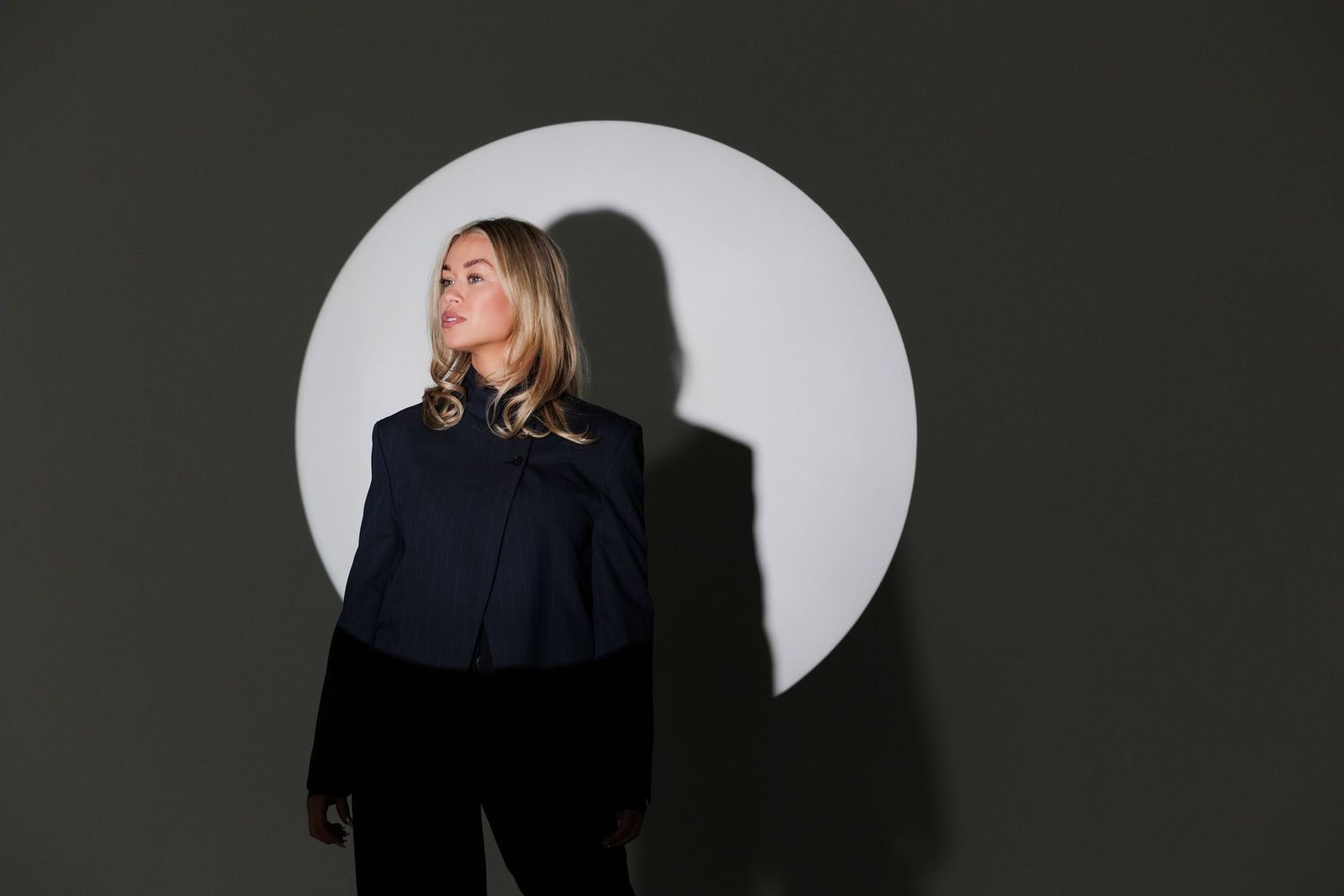 Blonde woman in navy blue top poses against dark wall with circular white light installation backdrop.