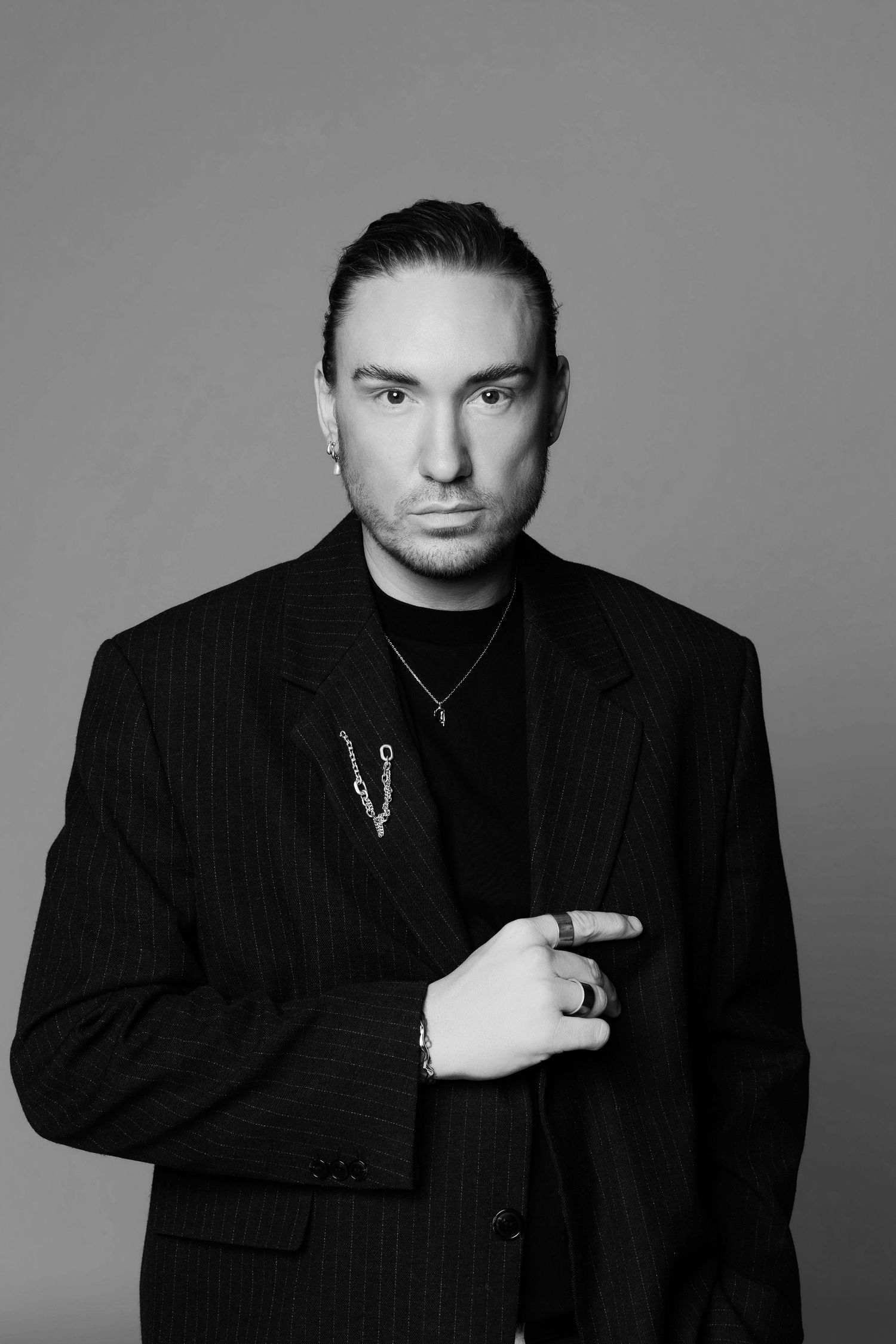 Black and white portrait of a man in a pinstripe suit wearing chain necklaces and rings, posing against a gray background.