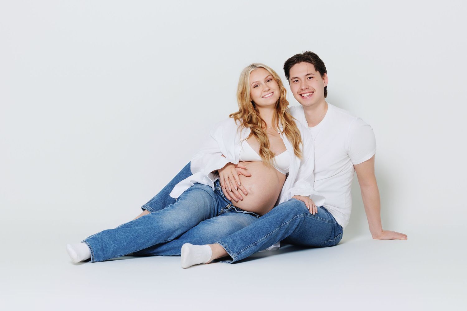 Smiling couple sitting on white floor, pregnant blonde woman and partner cradling her baby bump.
