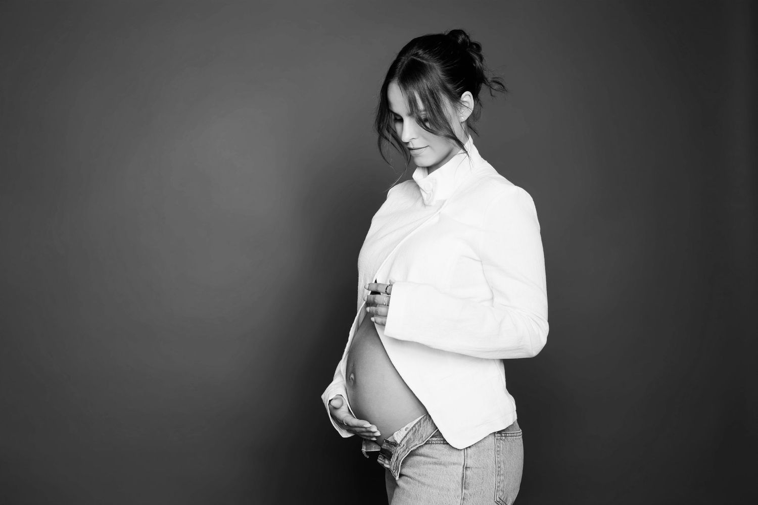Black and white photo of pregnant woman in white shirt and jeans gently holding her bare baby bump.