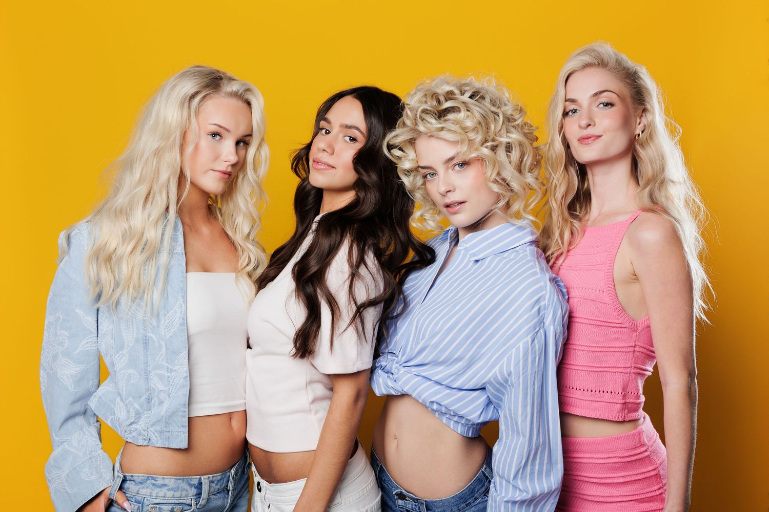 Four fashionable young women with blonde and dark hair pose together against a vibrant yellow background in trendy outfits.