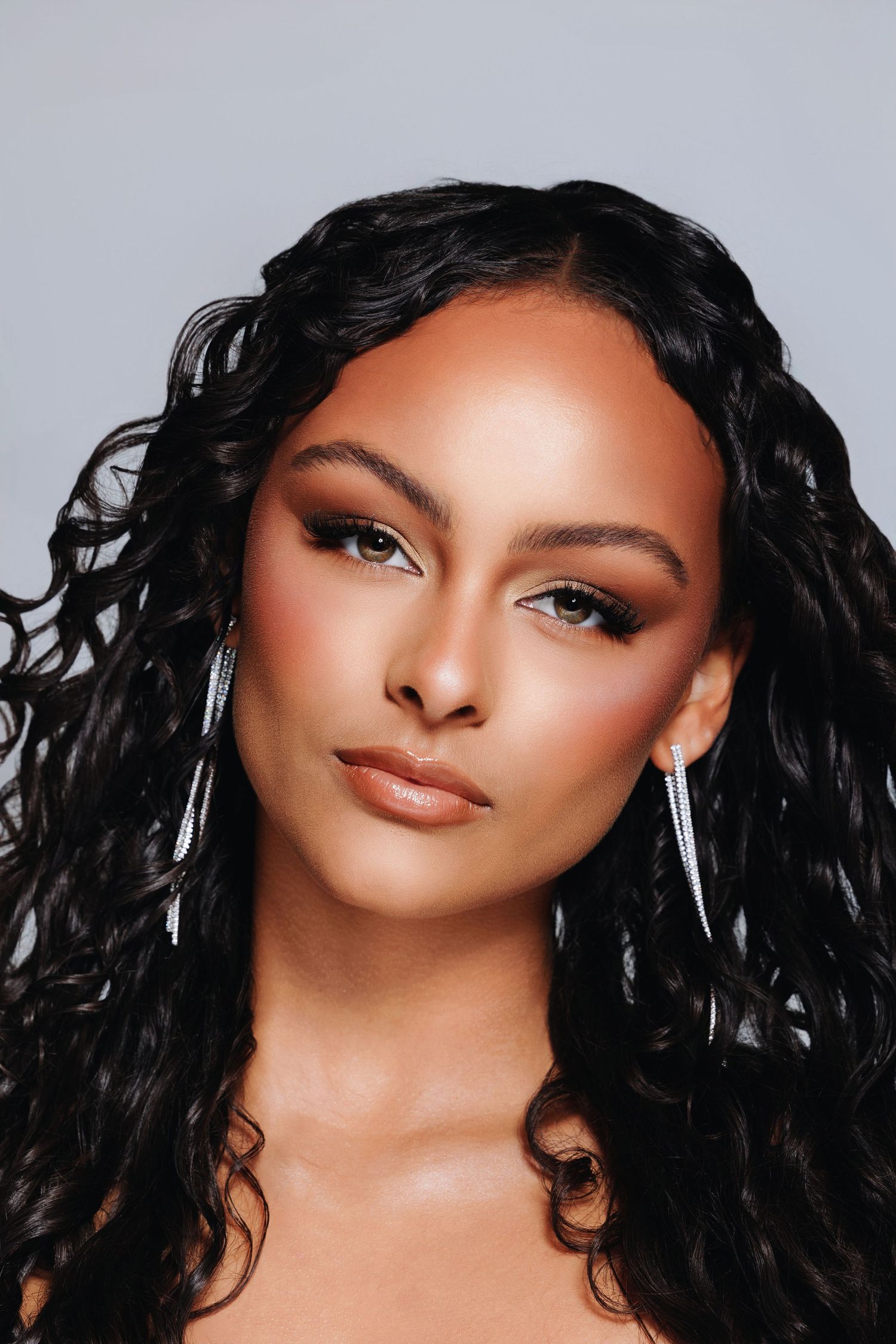 Young woman with long curly black hair, silver drop earrings, and glamorous makeup poses against a gray background.