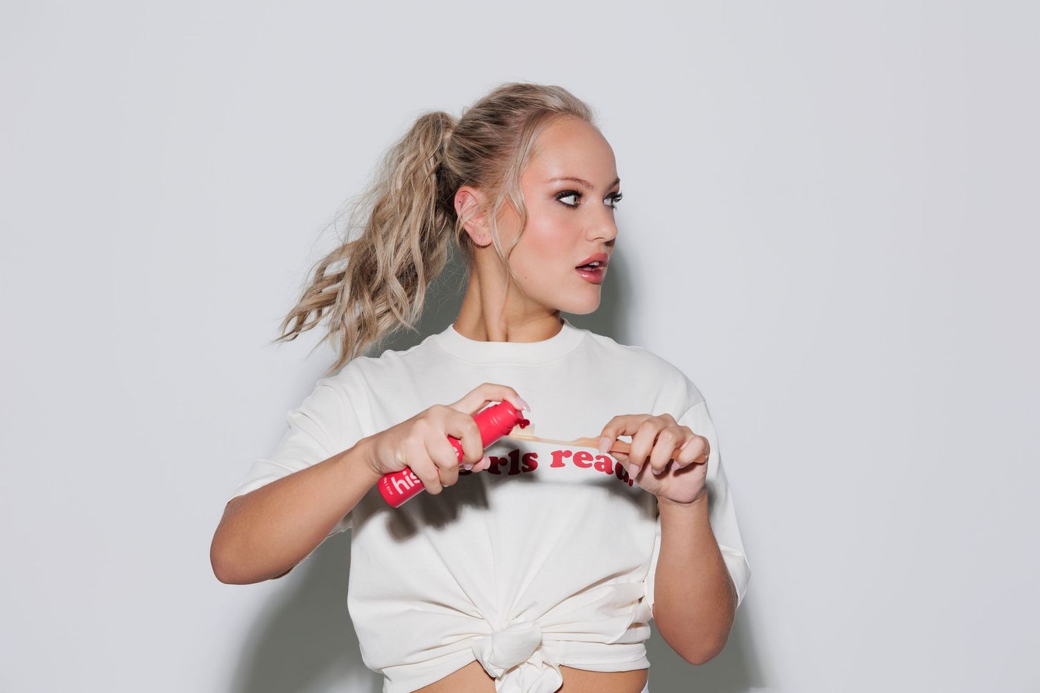 Young blonde woman in white crop tee holding red object, posing against light gray background with ponytail.
