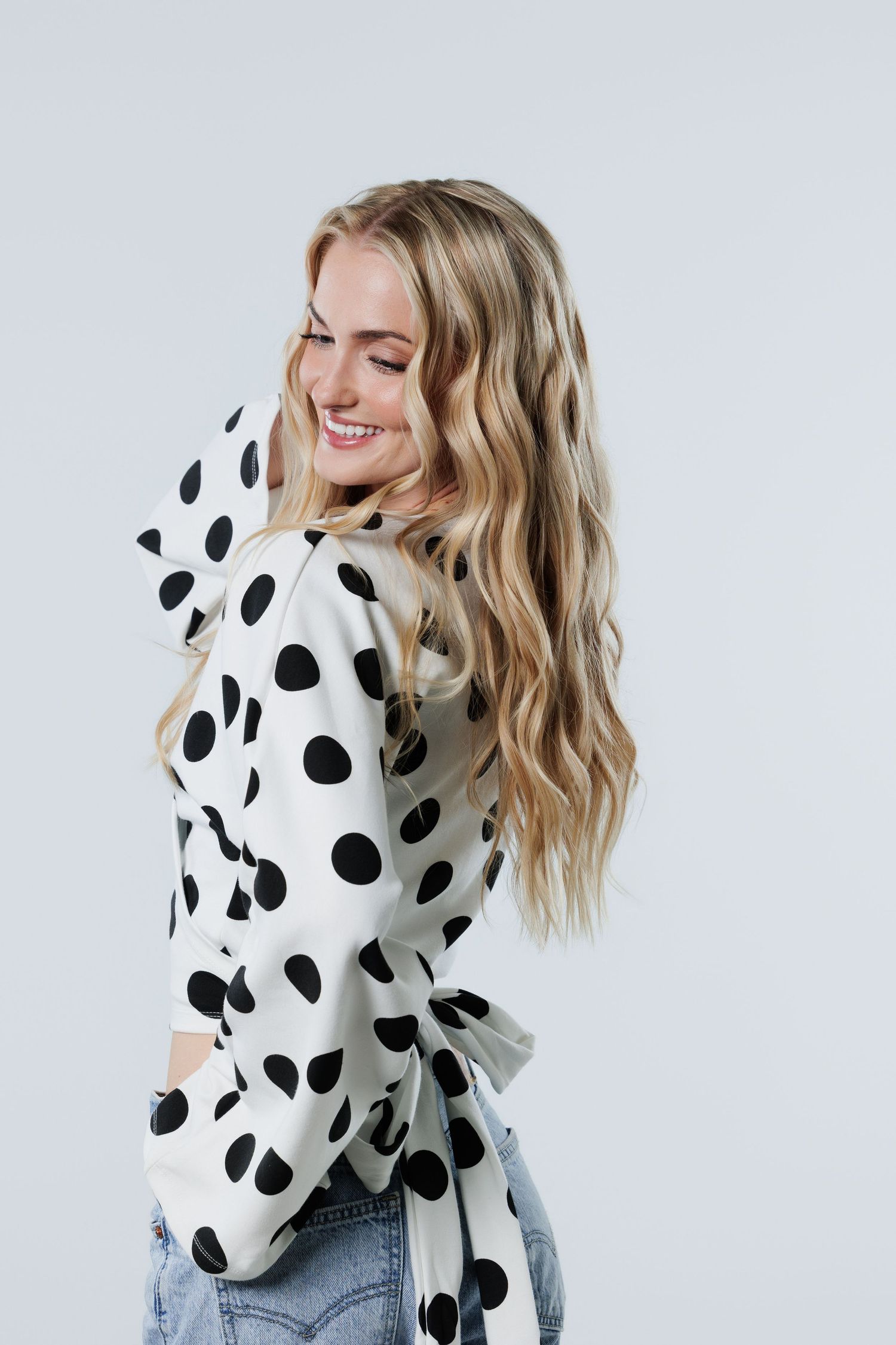 Happy blonde woman in polka dot blouse and jeans smiling over shoulder on grey background.