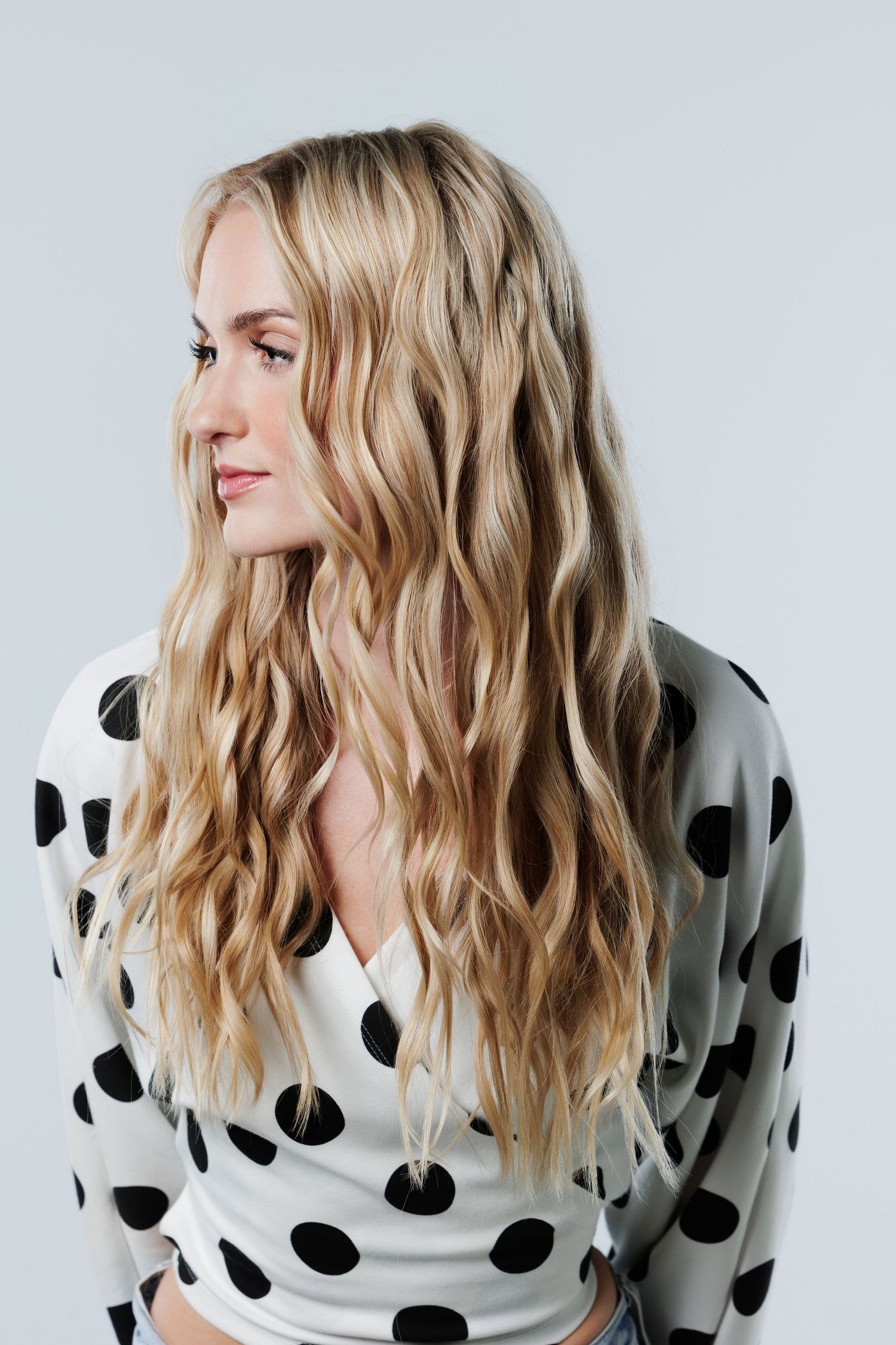 Young woman with long wavy blonde hair wearing a black and white polka dot top, posing against a light gray background.