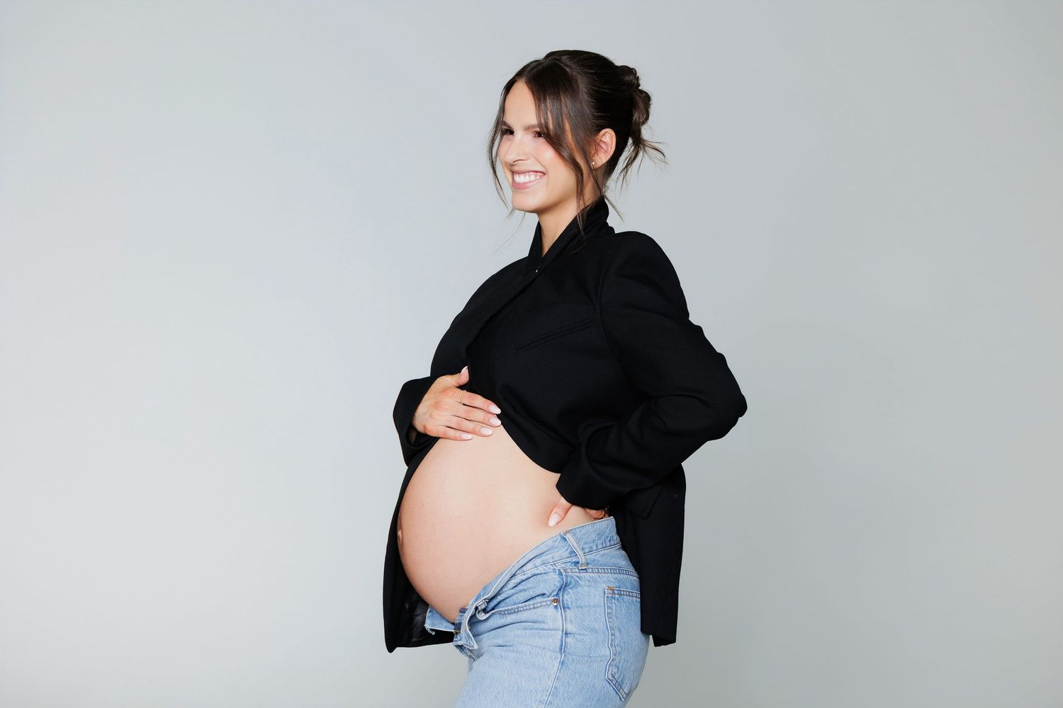 Smiling pregnant woman in black shirt and jeans posing sideways showing bare baby bump in studio.