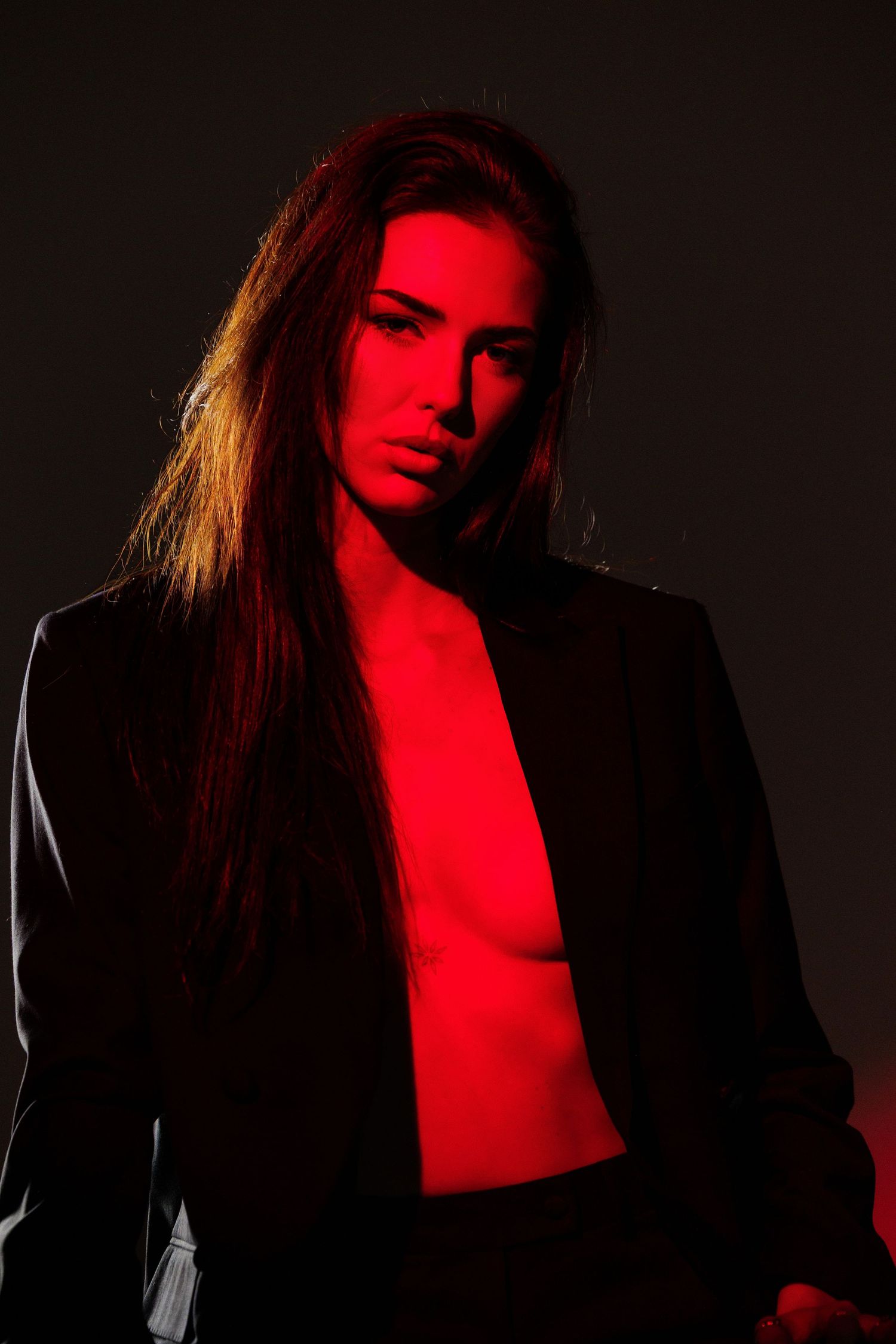 A woman with long hair poses in a black blazer under dramatic red lighting against a dark background.