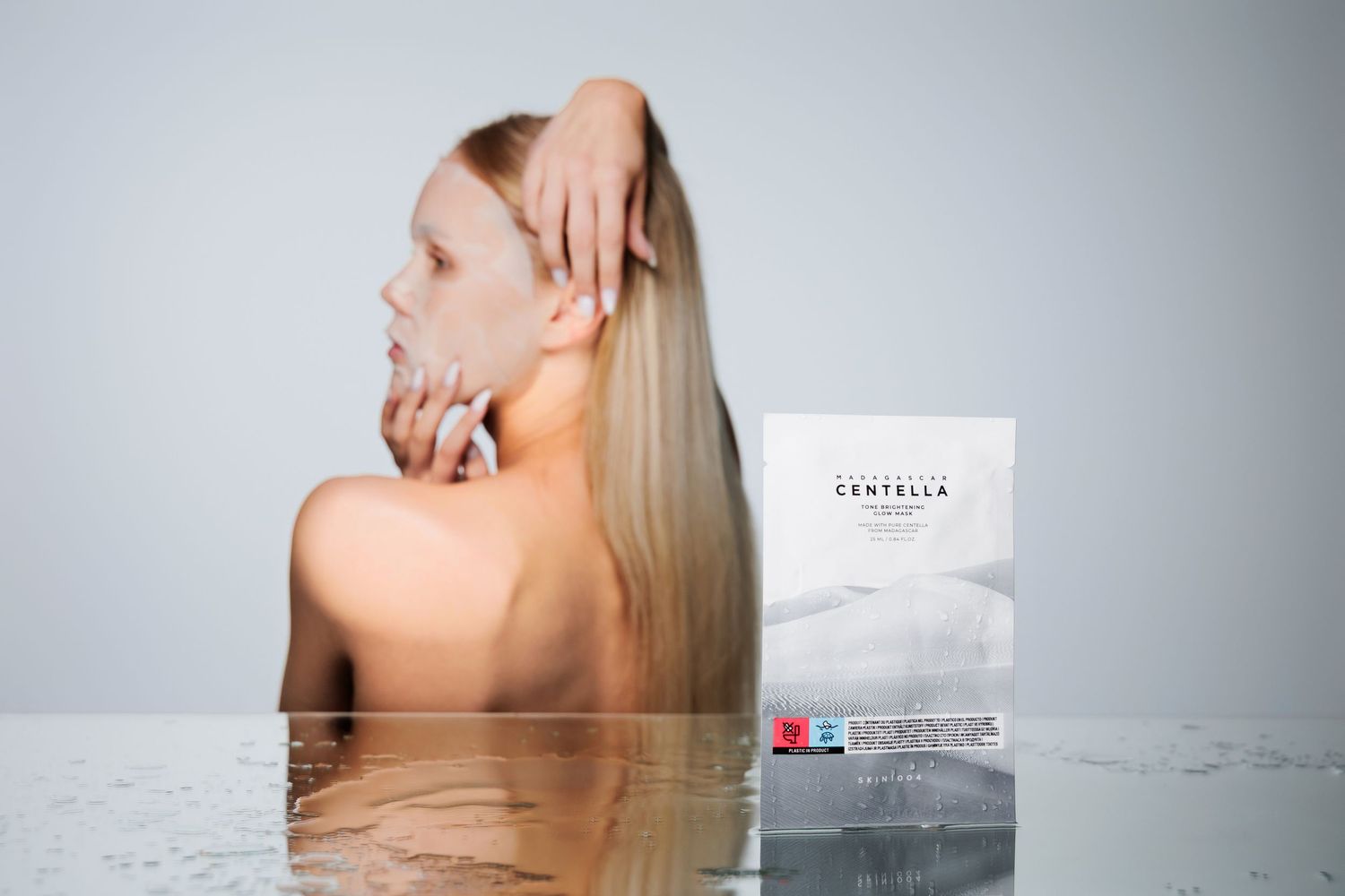 Woman with long blonde hair wearing a Centella face mask sheet, posed elegantly beside product packaging.